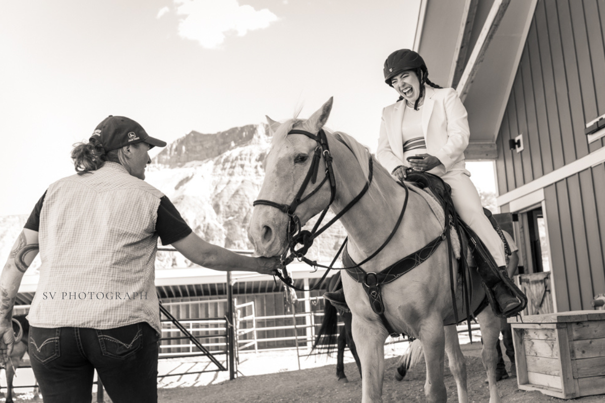 A rider sits on a light-colored horse as another person guides it near a stable with mountains behind.