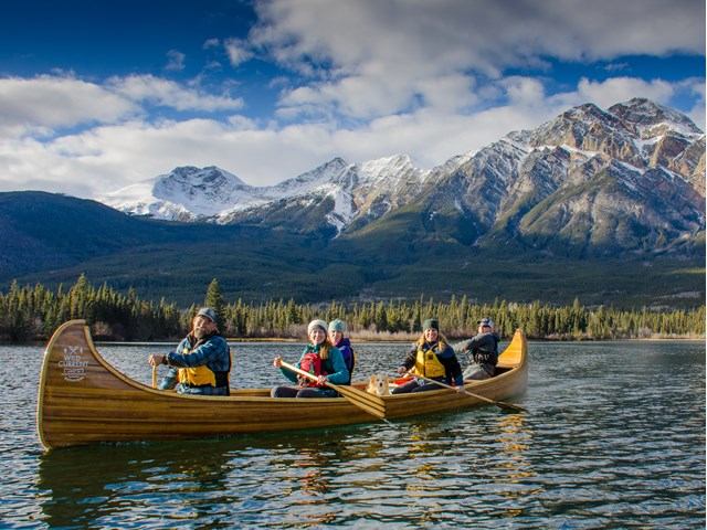 A group of five people canoeing on a calm lake in front of snow-capped mountains under a cloudy blue sky, exuding a sense of adventure and tranquility.