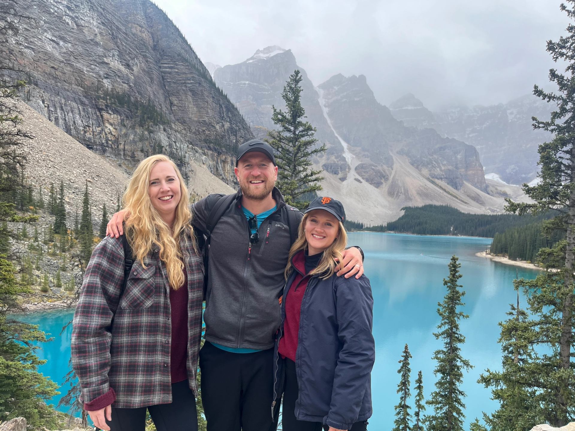 Three hikers stand by a turquoise mountain lake with rocky peaks and trees behind.