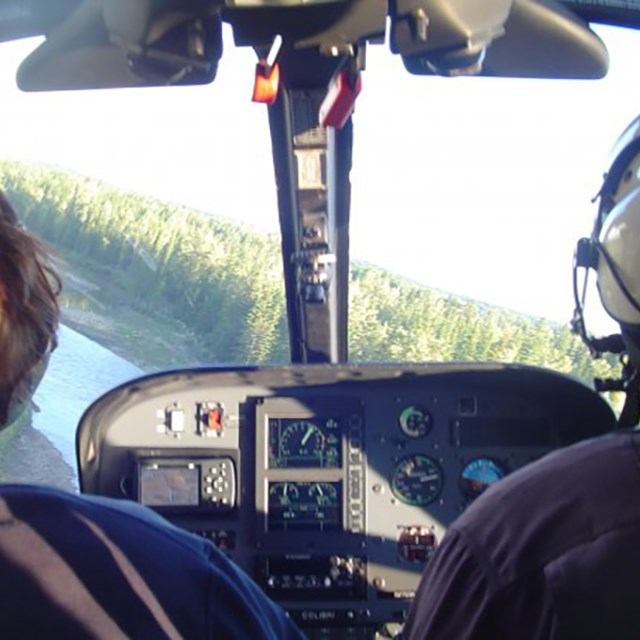 Helicopter cockpit with instrument panel and view of river and forested hills.