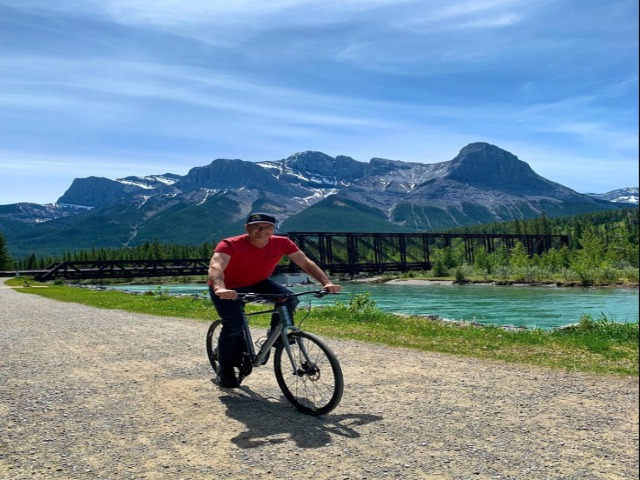 Cyclist on gravel path with river, bridge, and mountain backdrop under clear sky.