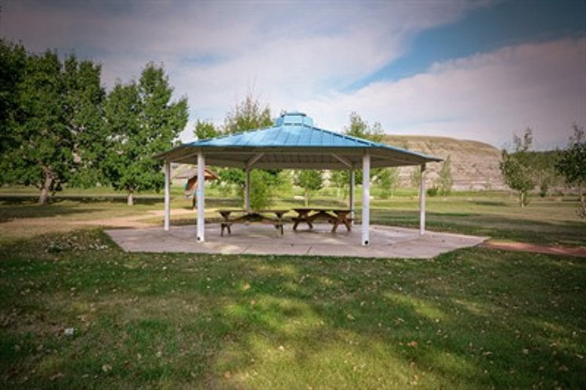 Gazebo with picnic tables in grassy area near hills under clear sky.