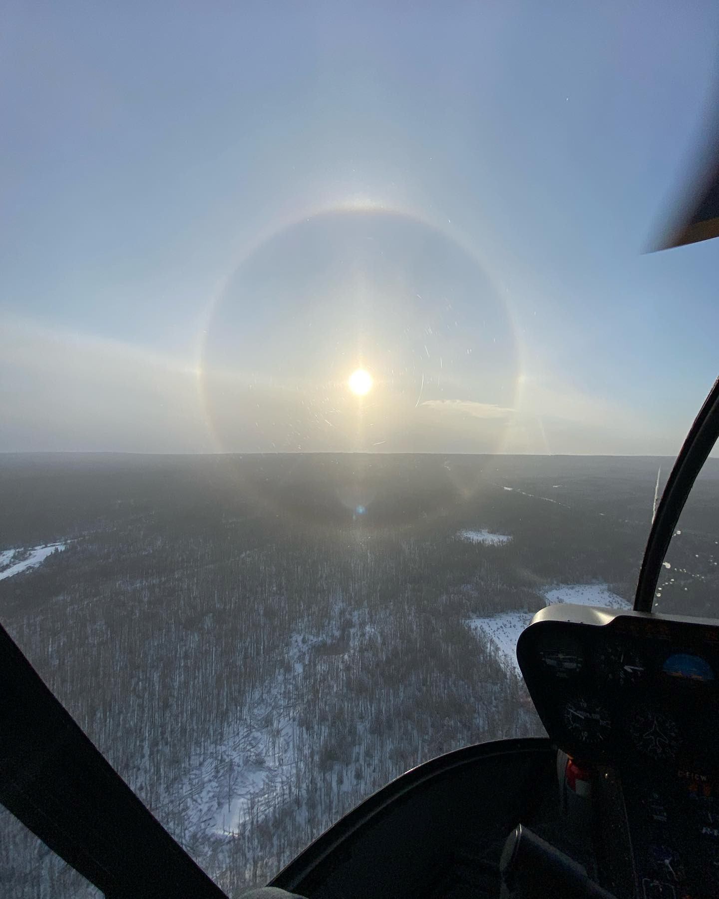View from helicopter cockpit over snowy forest with sun halo.