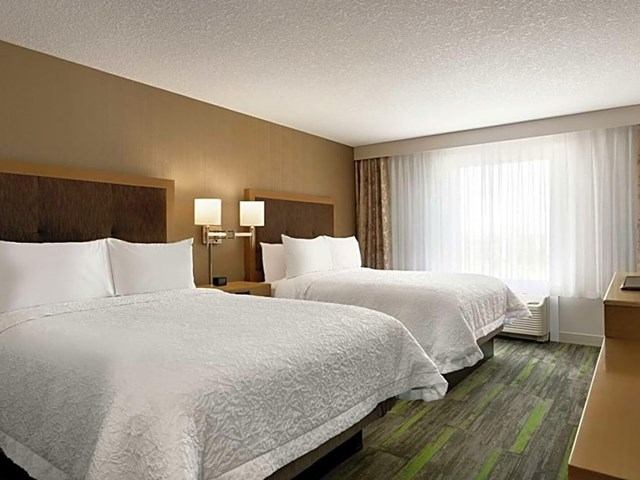 Hotel room at The Landing with two beds, wall lamps, and large window with sheer curtains.