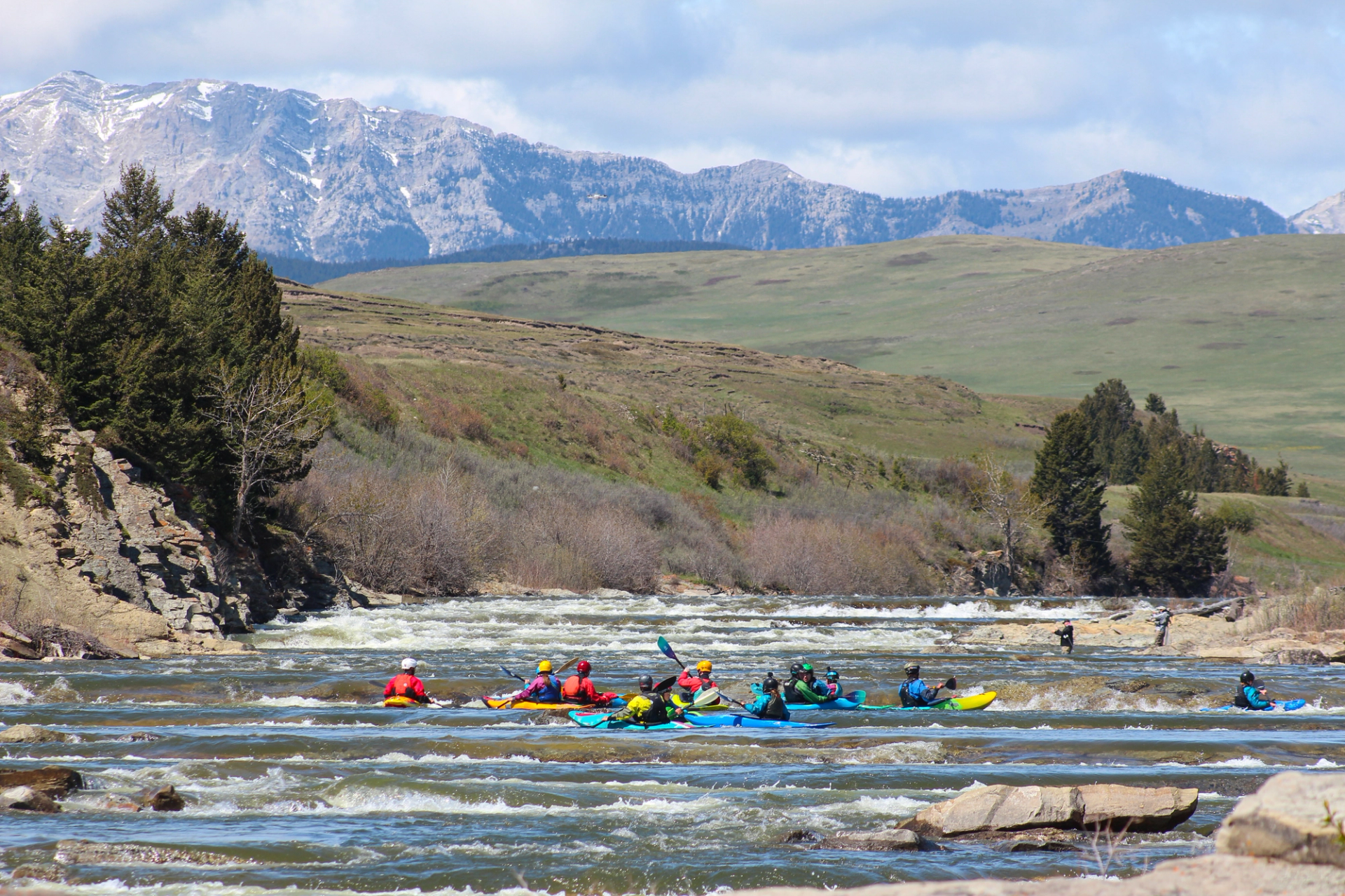 Group of kayakers paddling through river rapids with mountains in the distance.