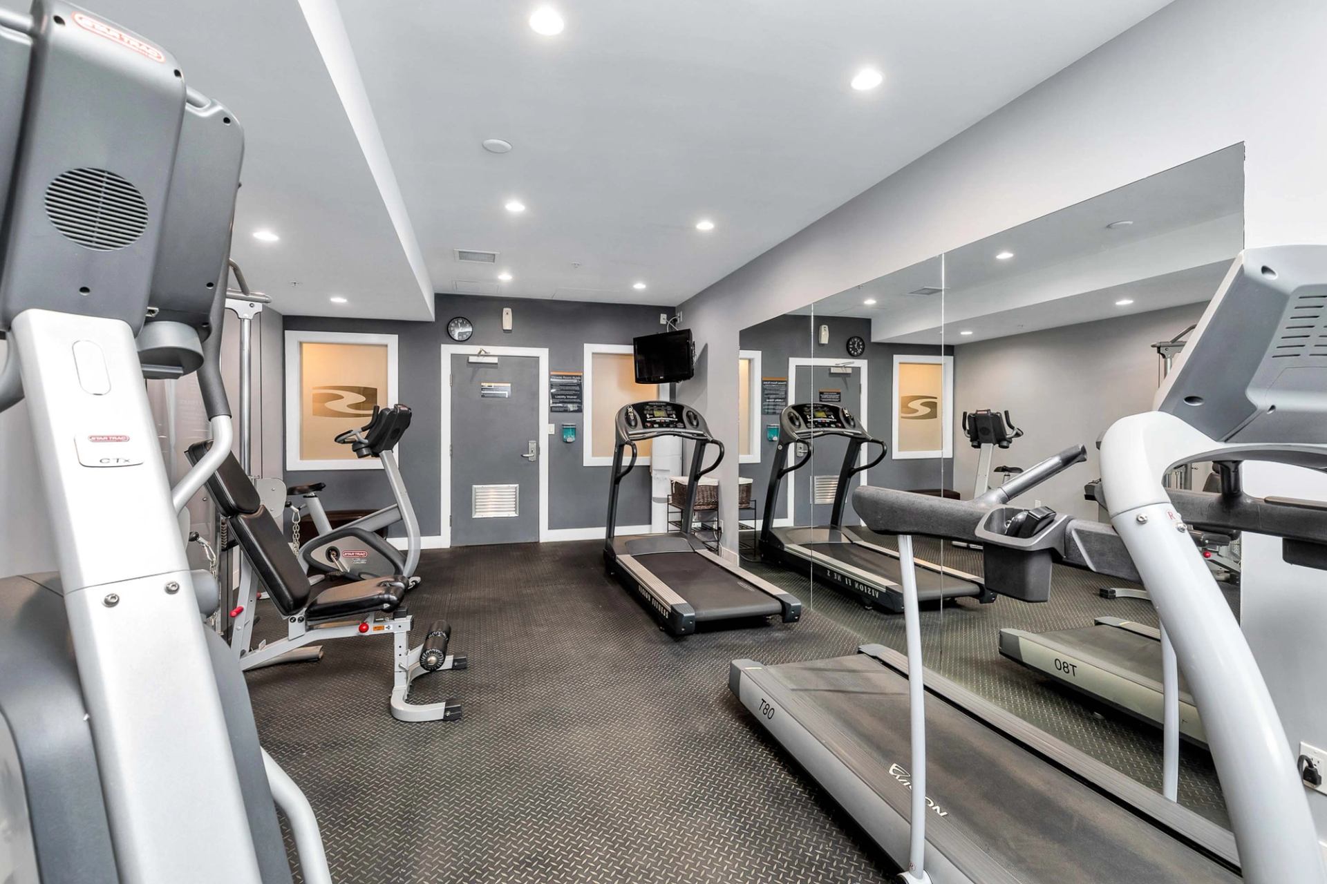 Fitness room with treadmills, stationary bikes and weight machines facing mirrored wall and doors.