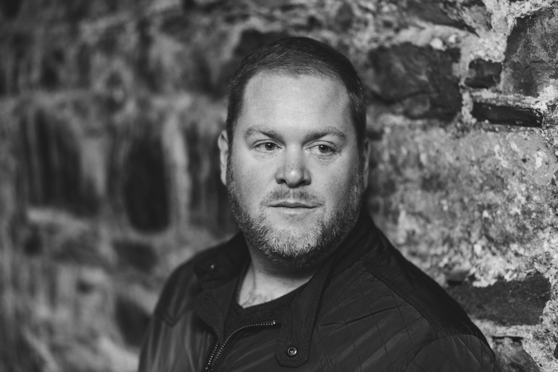 Man with short hair and beard stands by rough stone wall, looking sideways.