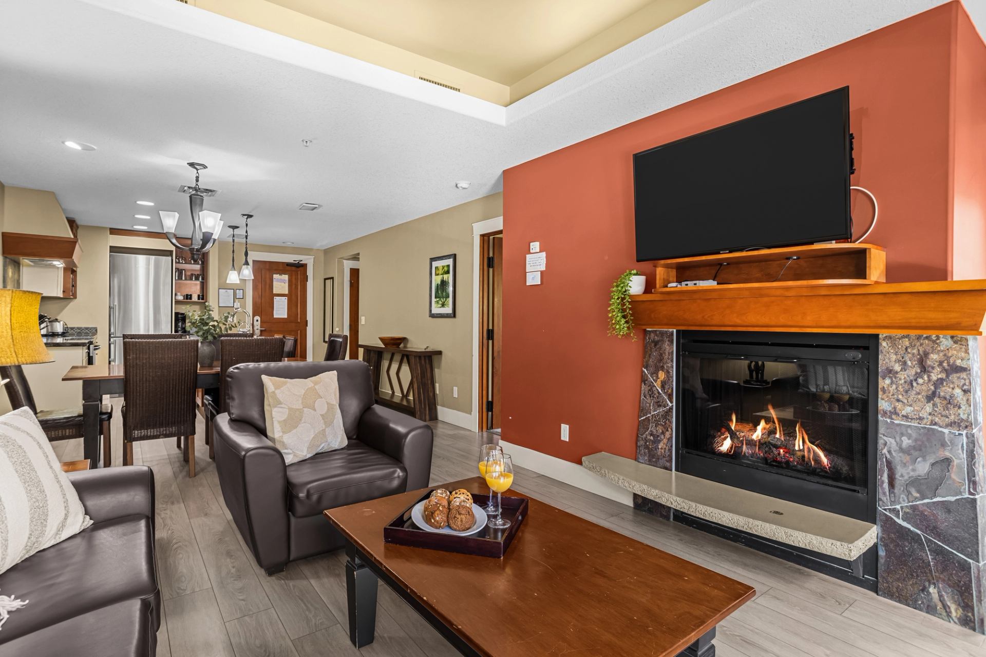 Living room with leather seating, a lit fireplace, wall-mounted TV and adjoining kitchen.