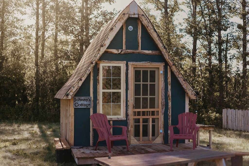 A quaint wooden cottage labeled "Johnny's Cottage" stands in a sunlit clearing. Two red chairs are on the front porch, surrounded by tall trees. Cozy and inviting atmosphere.