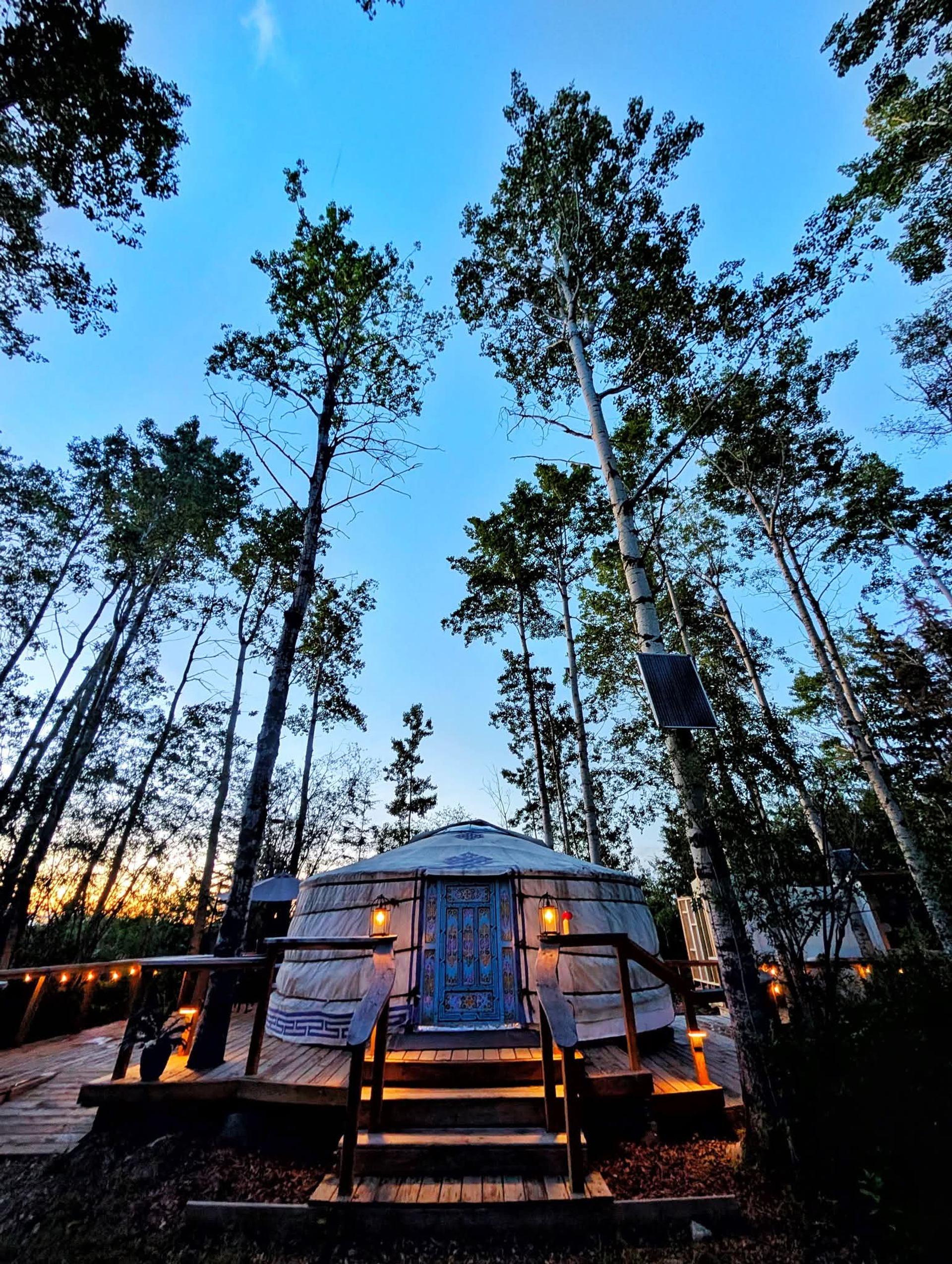 An illuminated yurt with an ornate blue door on a wooden deck, surrounded by tall trees at dusk.