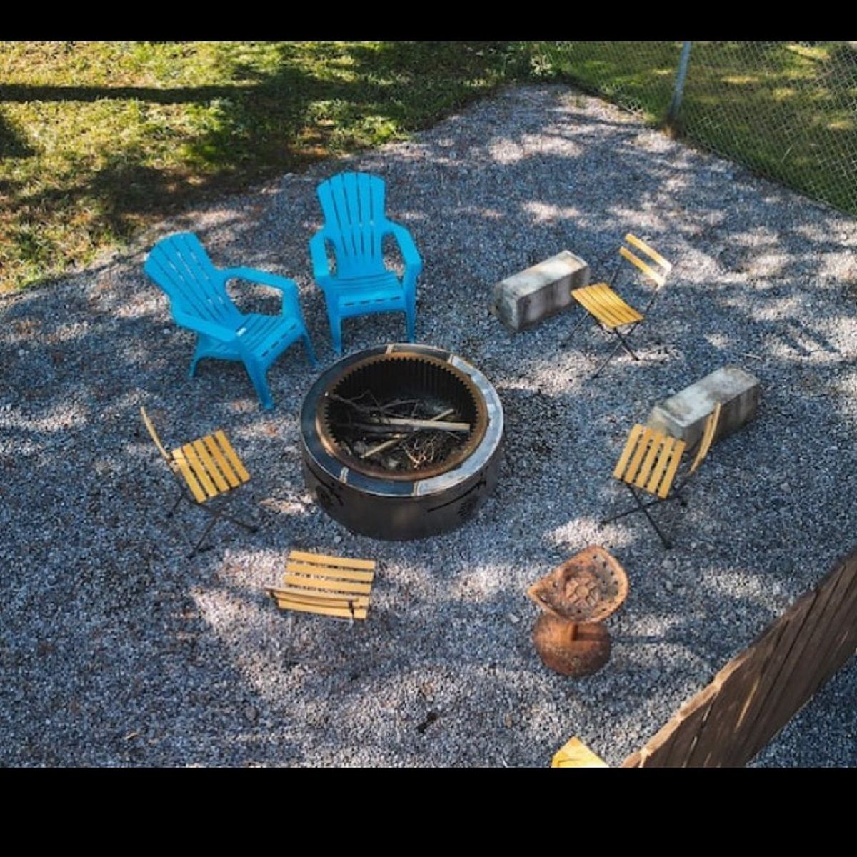 Outdoor firepit surrounded by blue chairs and small wooden seats on a gravel patio.