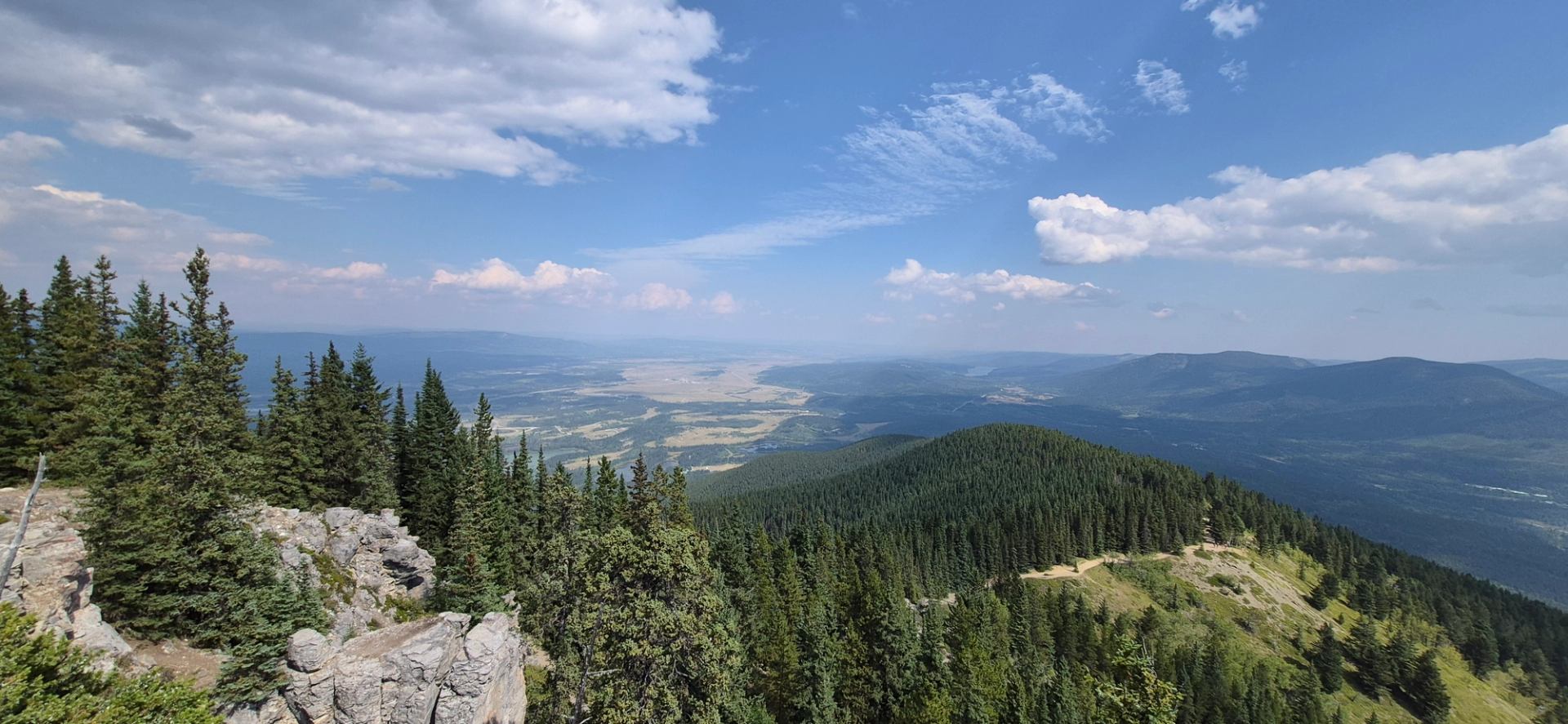 Rocky mountain ridge overlooks evergreen forests and a broad valley beneath scattered clouds.
