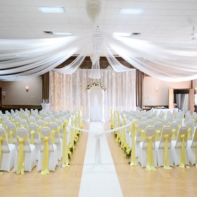Event hall with white aisle runner, chairs with yellow sashes, draped ceiling and arch.