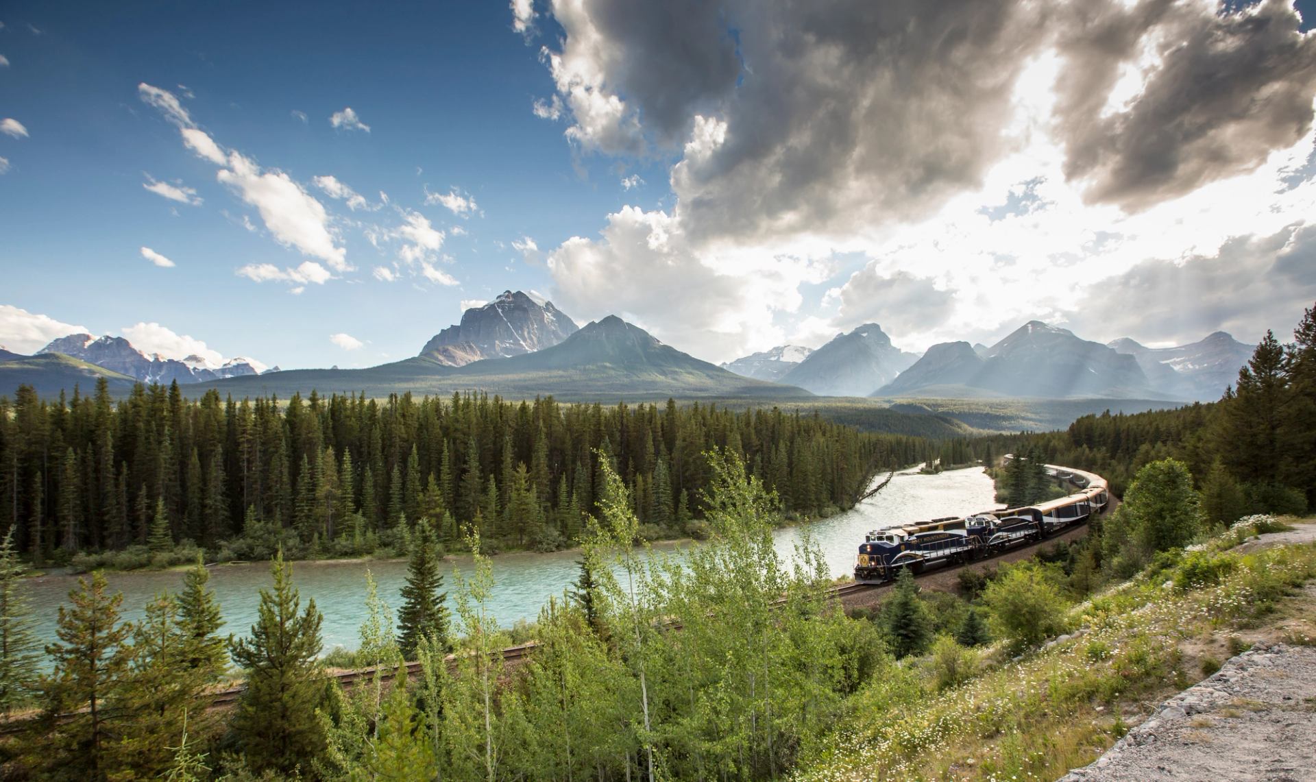 Rocky Mountaineer train traveling beside a turquoise river through forested valleys and mountain peaks.
