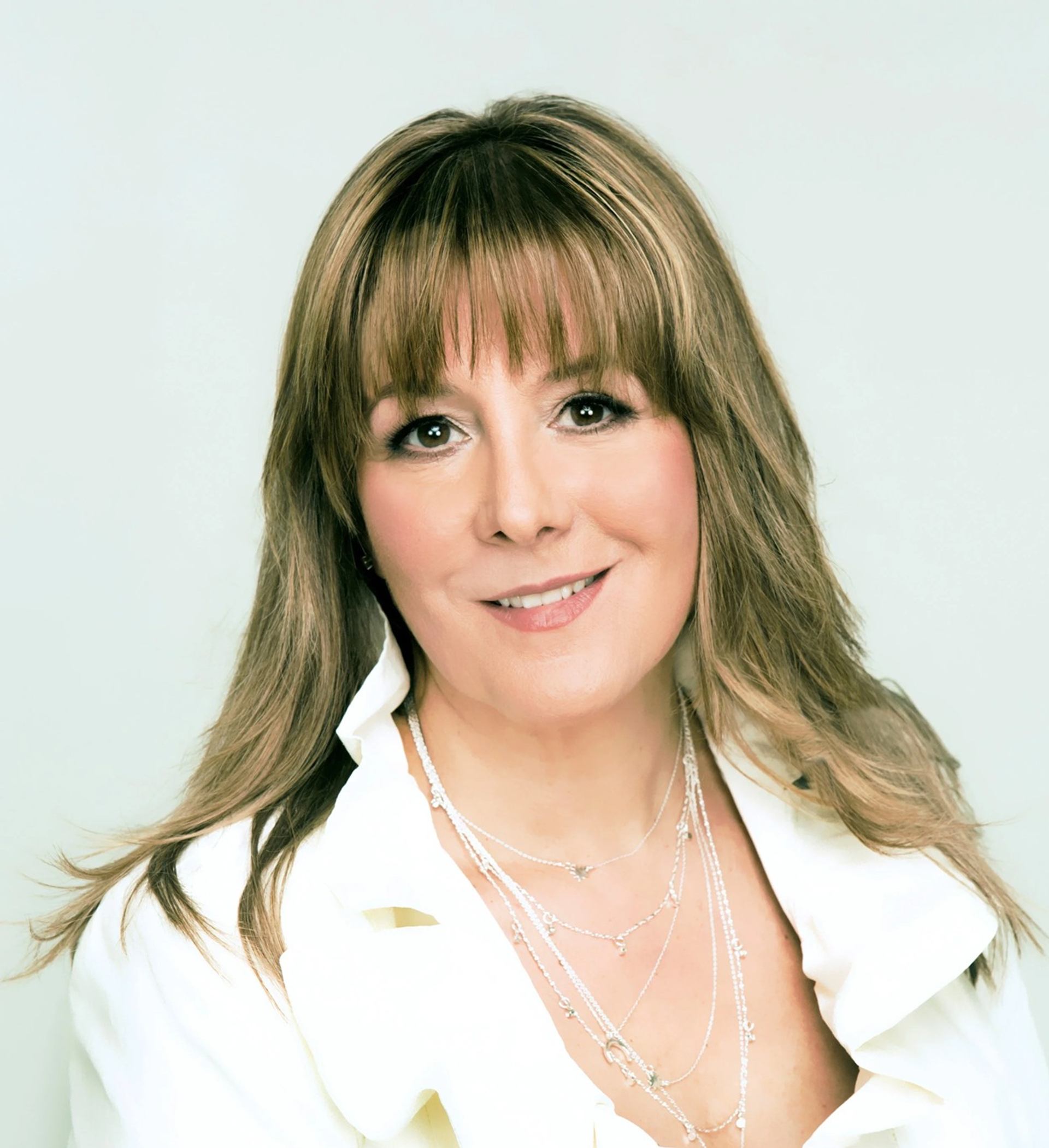 Person with long light brown hair in white blouse and layered necklaces, smiling.