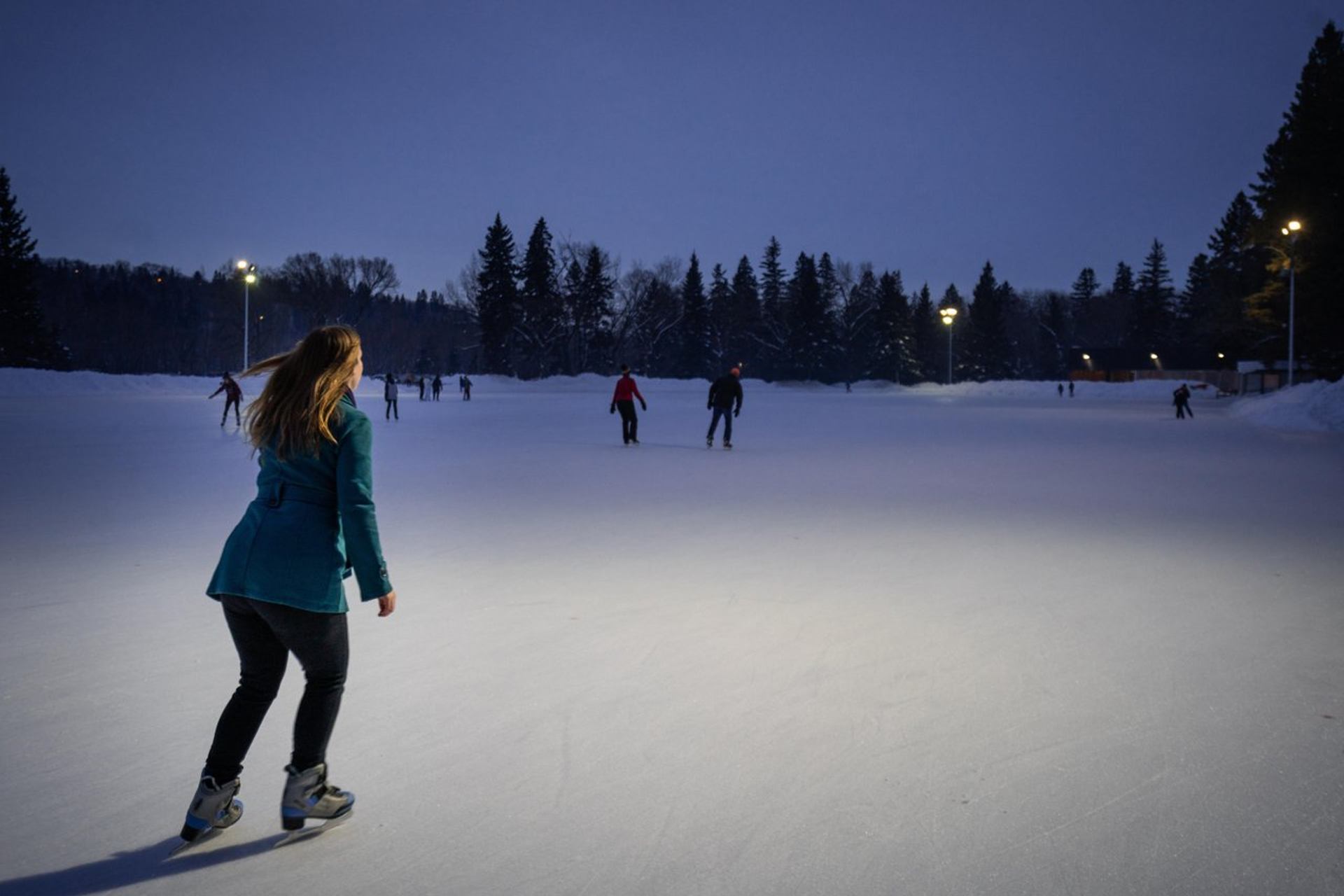 People skate on an outdoor rink at dusk, surrounded by snow and trees.
