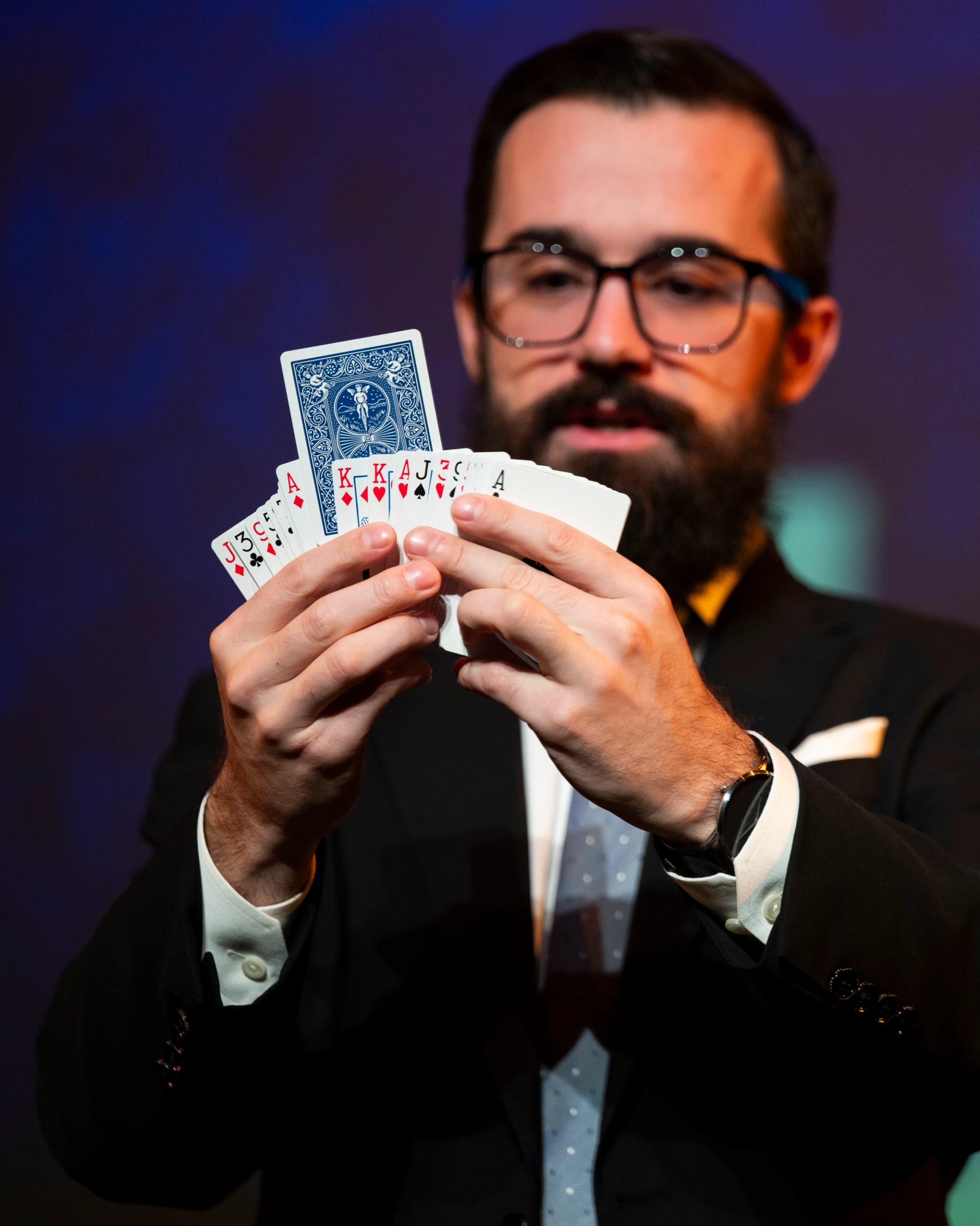 Magician in suit holding fanned playing cards, including hearts and a blue-backed card.