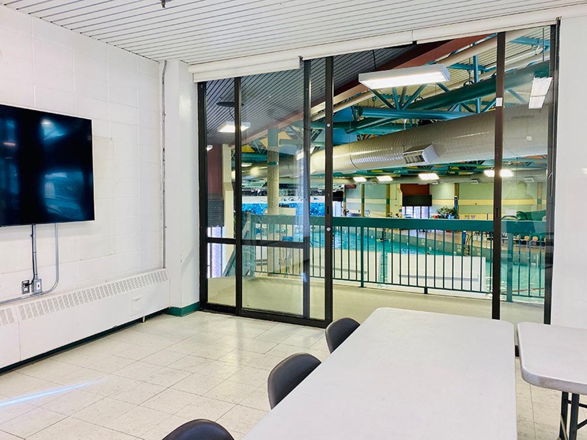Meeting room with tables, wall-mounted TV and window overlooking indoor swimming pool.