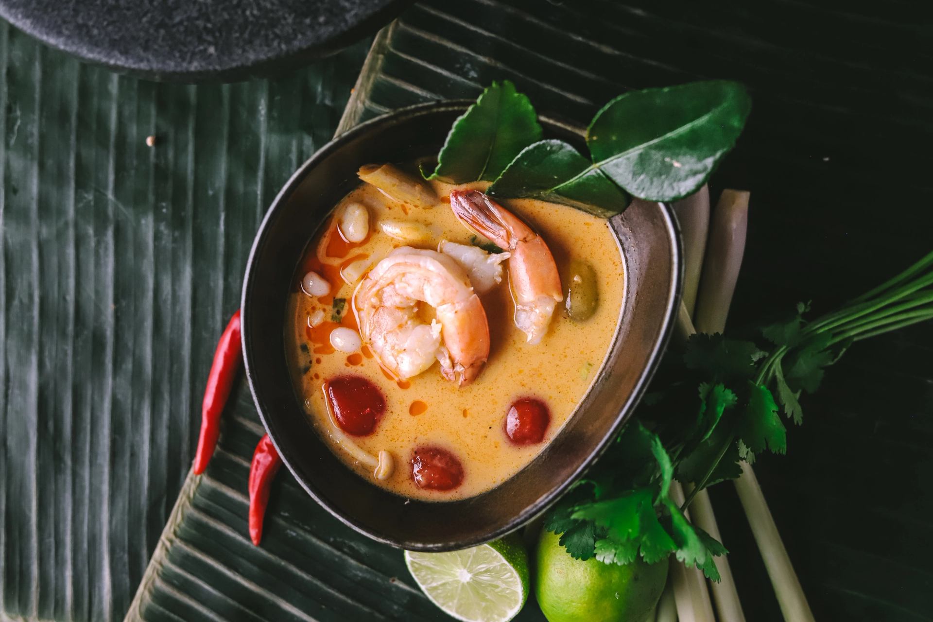 Bowl of shrimp soup with chilies, lime, lemongrass and herbs on banana leaf.