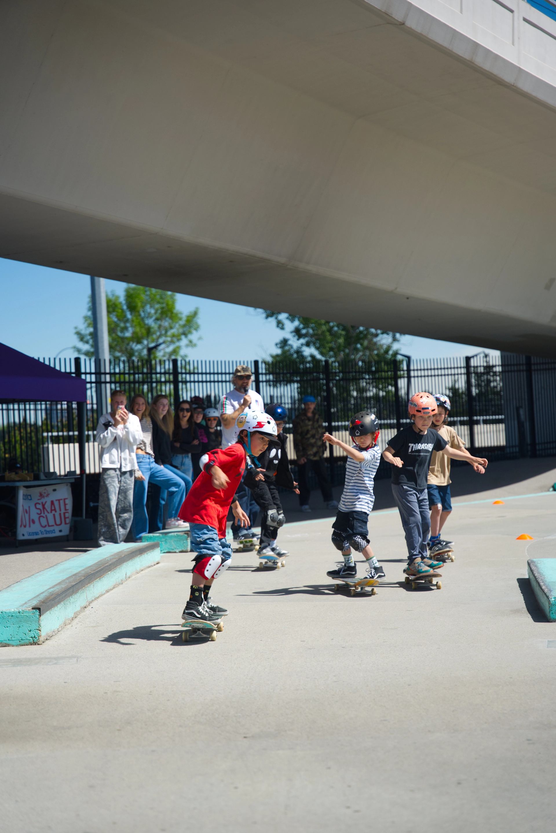 Children wearing helmets skateboard in a concrete park while onlookers watch near a tent.