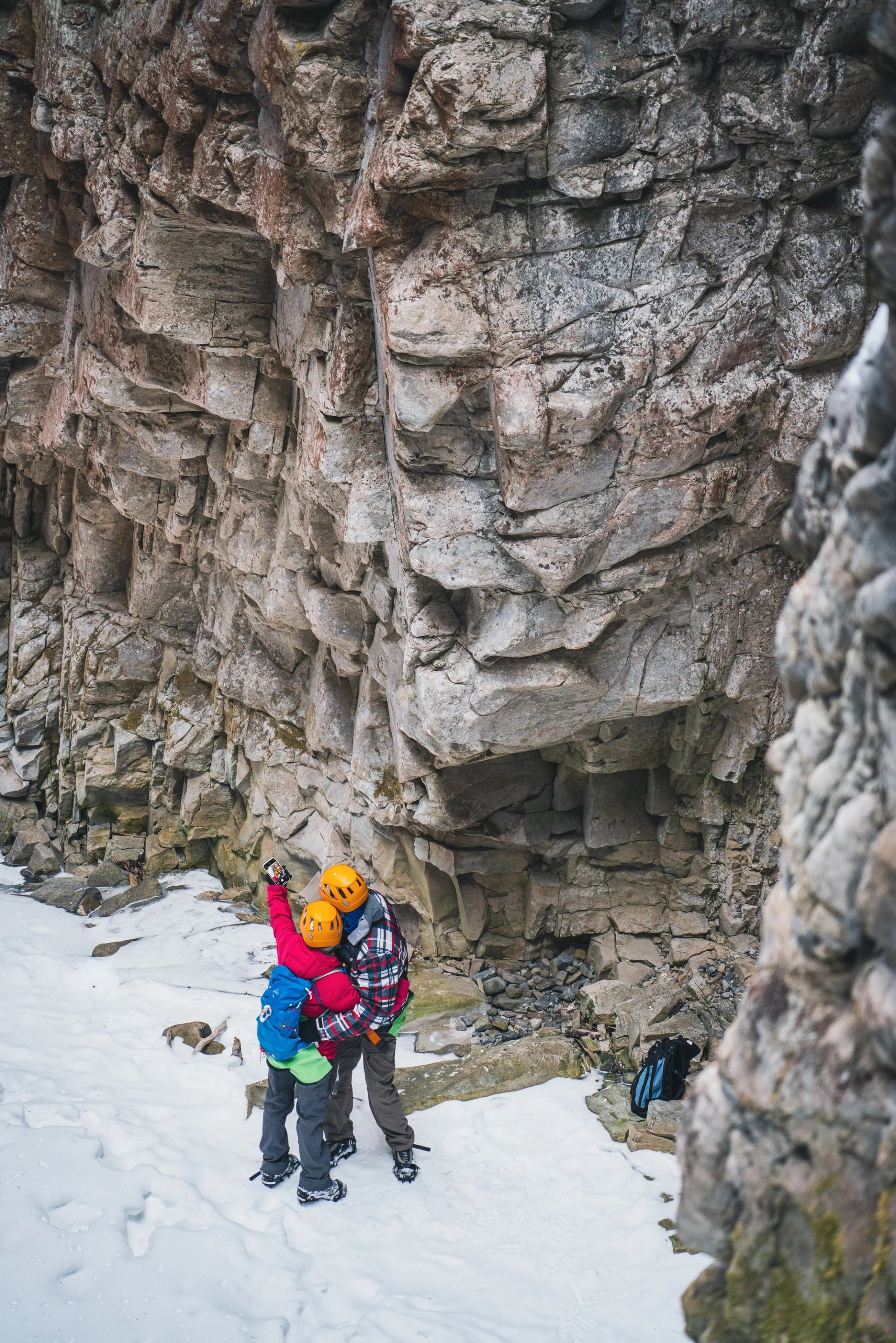 Two people in helmets and winter clothing stand by a rocky canyon wall in snow.
