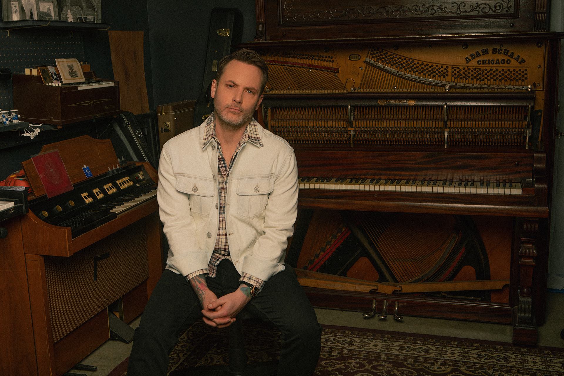 A man in a white jacket and plaid shirt sits between an organ and an antique "Adam Schaaf Chicago" upright piano with its internal mechanisms exposed.