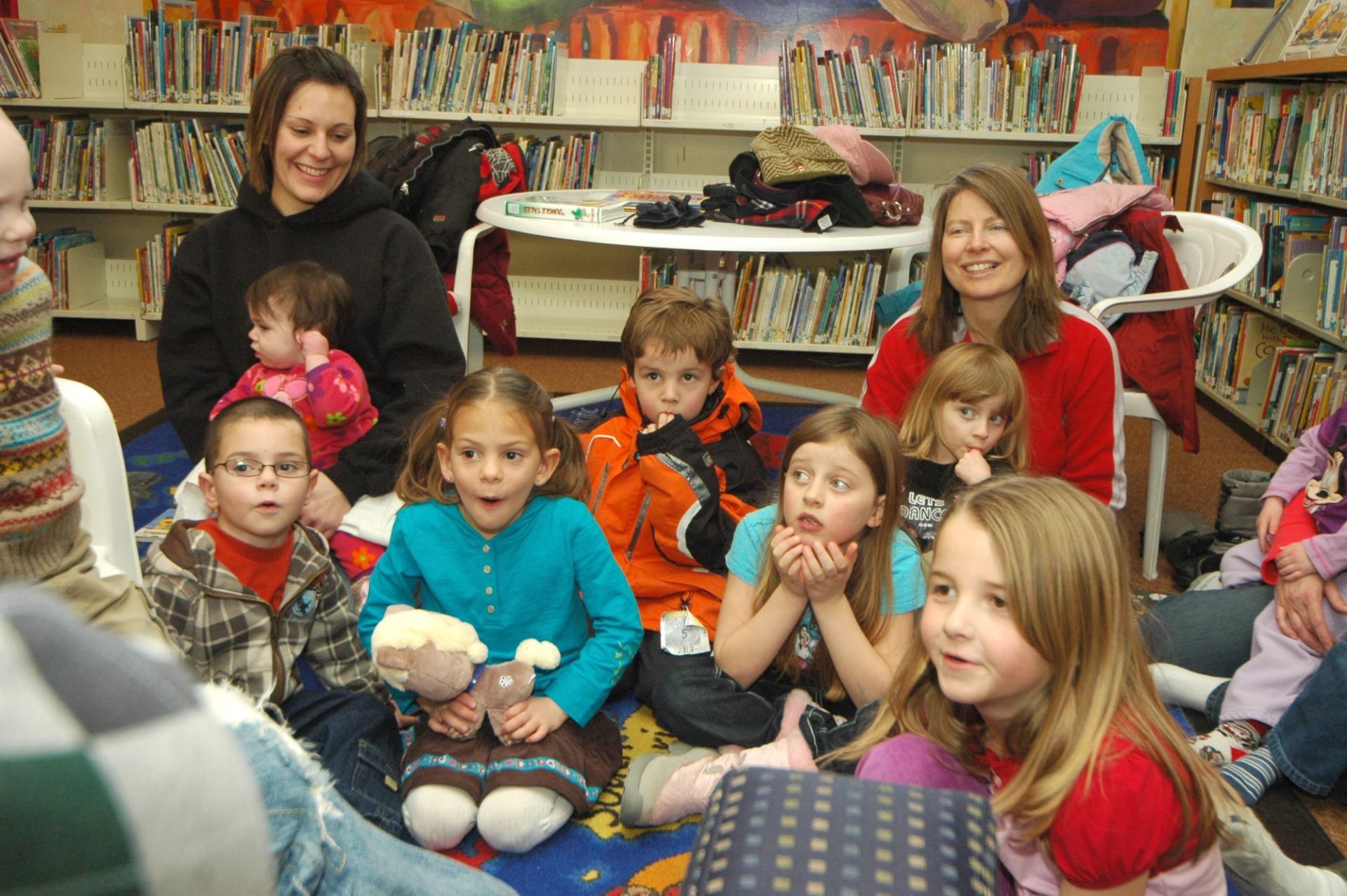 Children and adults sit on rug in library, bookshelves behind them.