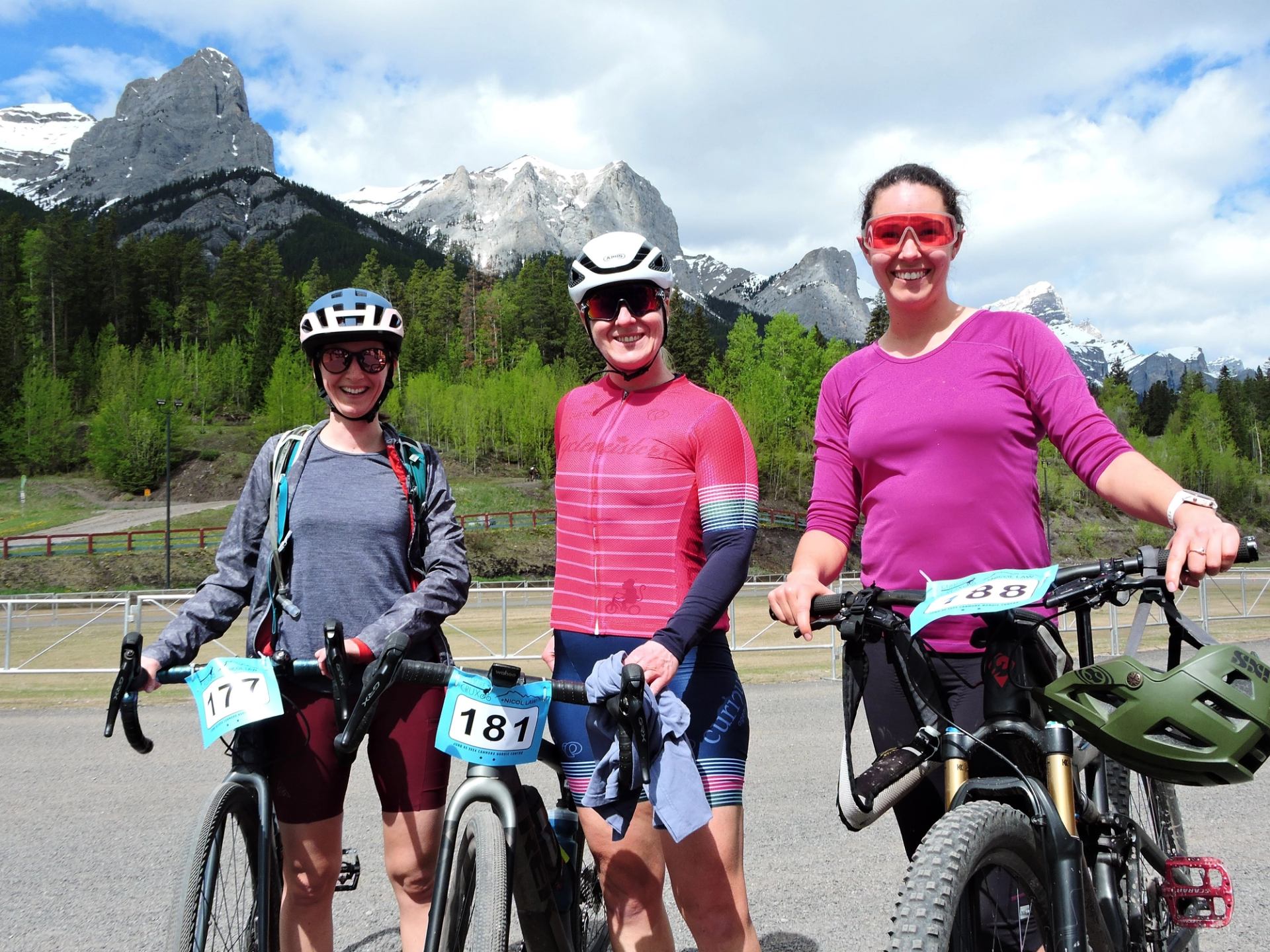 Three smiling women in cycling gear and helmets stand with their bikes (two gravel, one mountain bike) against a backdrop of mountains and a forest.