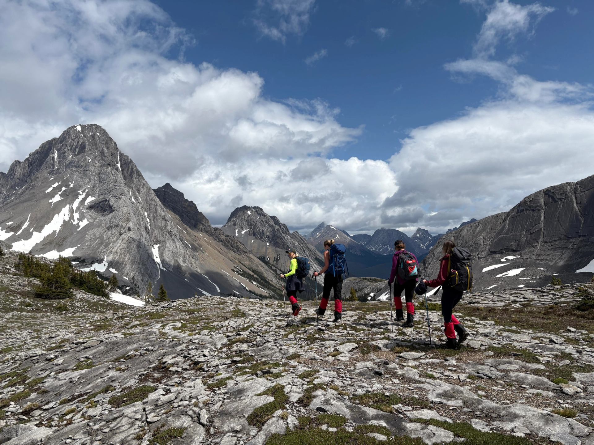 Group of hikers on rocky terrain with dramatic mountain peaks under a partly cloudy sky.