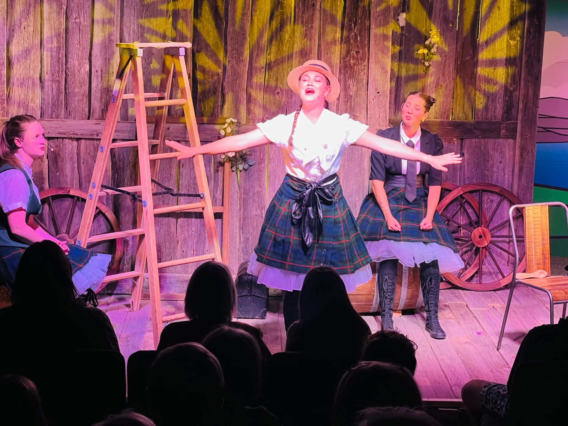 Performer sings center stage with arms outstretched; rustic set and seated cast behind.