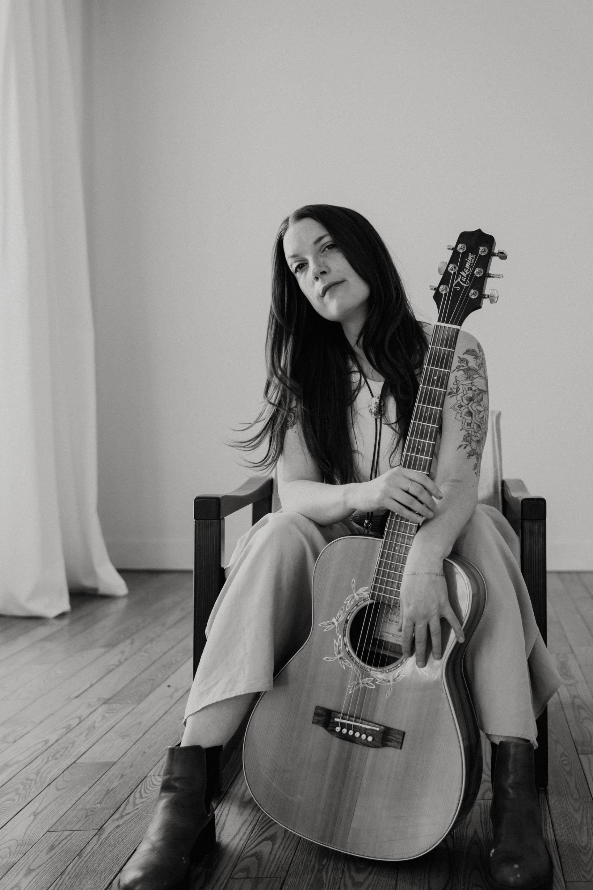 Person seated on wooden chair holding acoustic guitar in minimalist room.