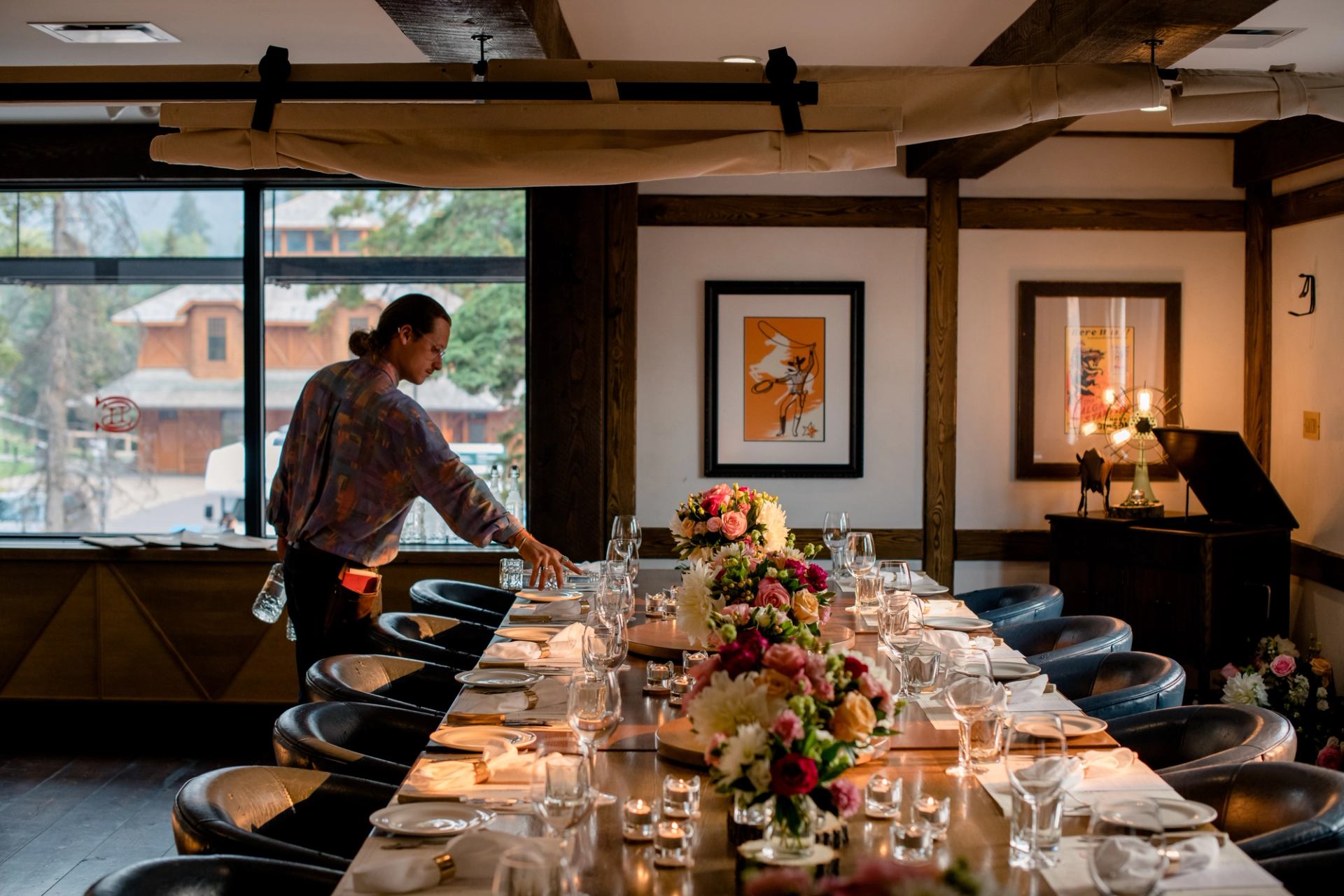 A staff member sets a long dining table with colorful floral centerpieces in a restaurant with large windows.