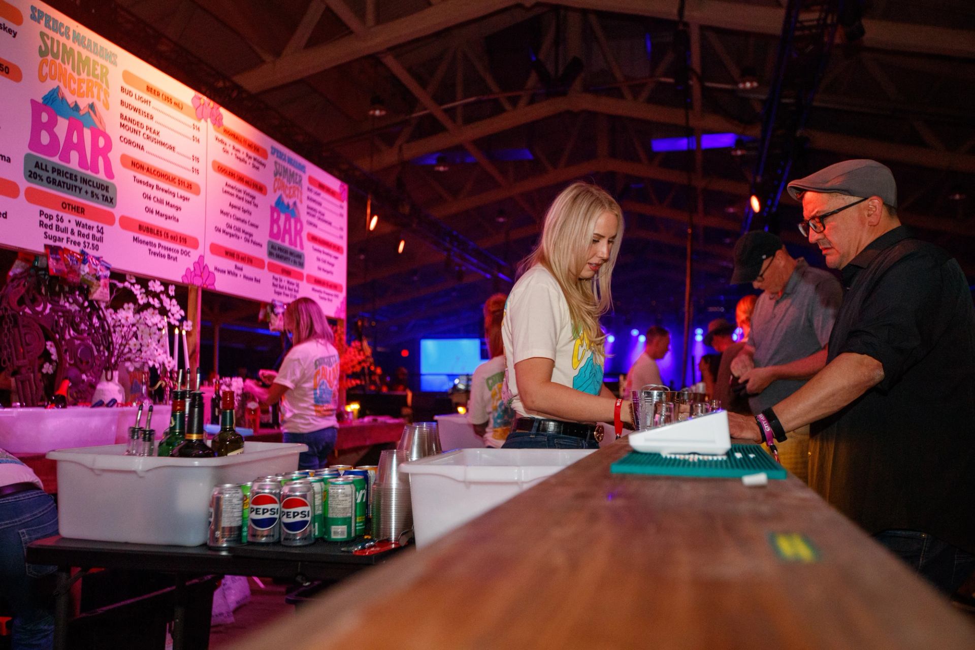 Bartender serves drinks at a concert bar with bright menu signs overhead.