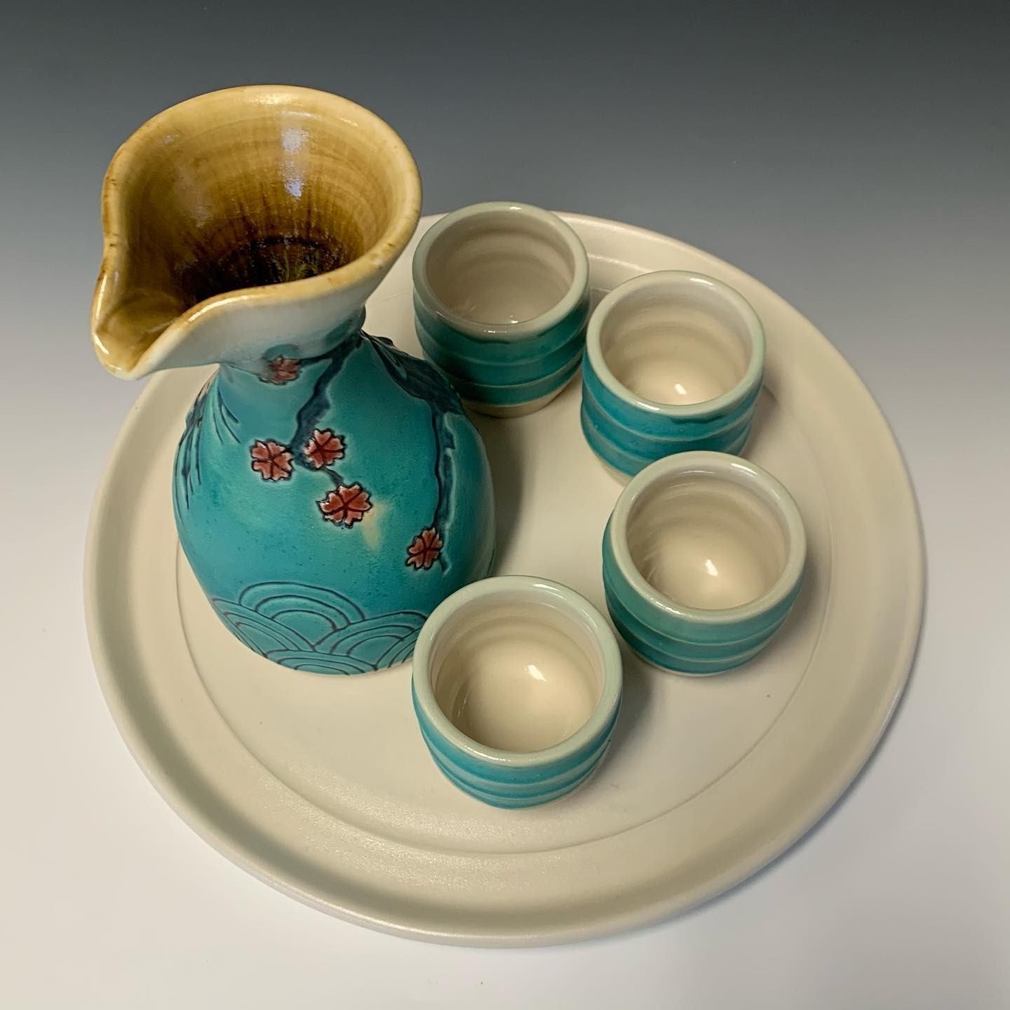 Turquoise ceramic pitcher with spout and small cups on a white platter.