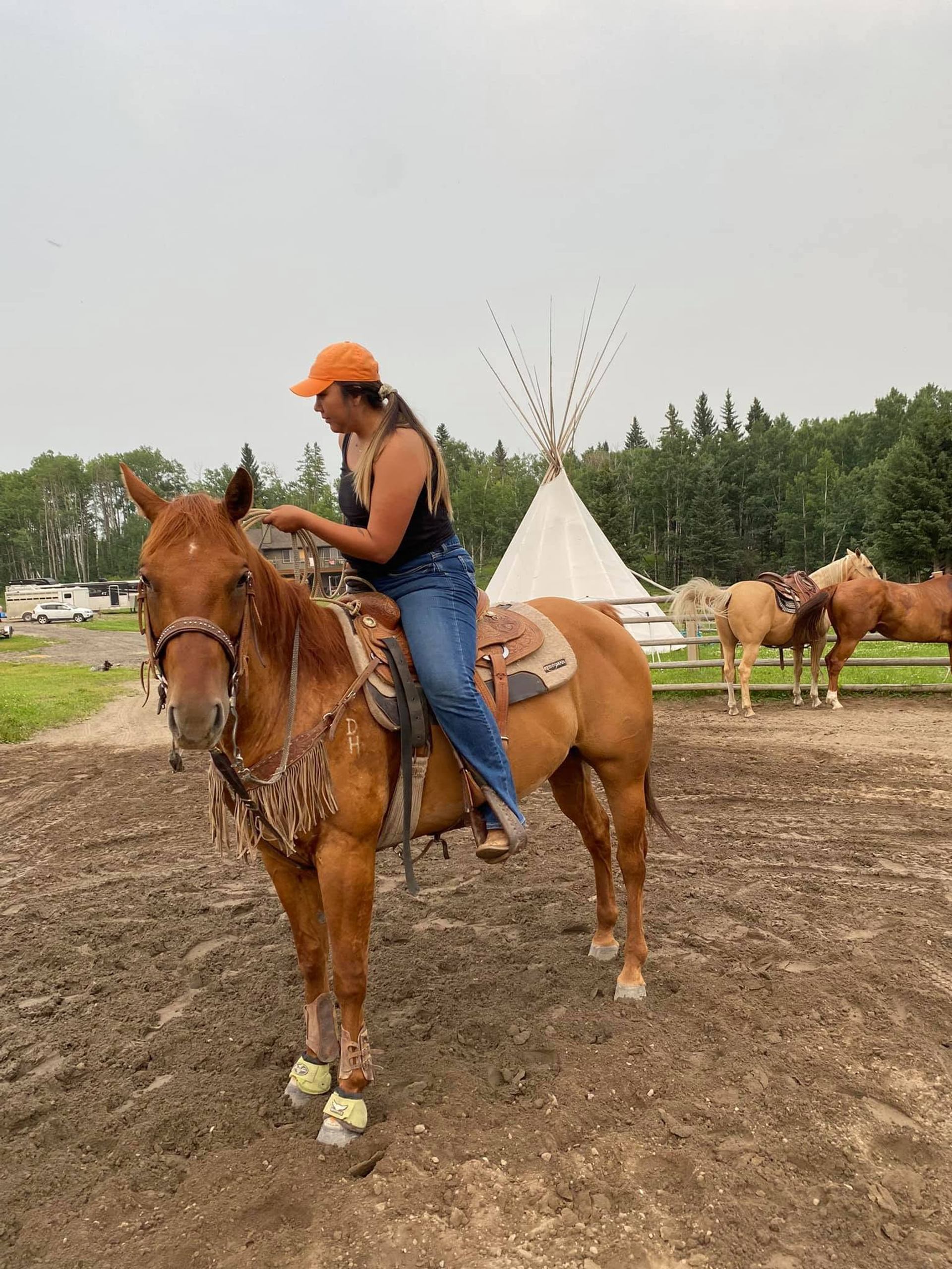 Person on saddled horse in dirt corral with teepee and trees in background.