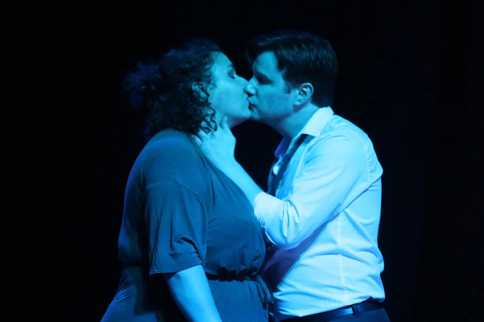 Two performers on stage in an intimate pose under dramatic blue lighting.
