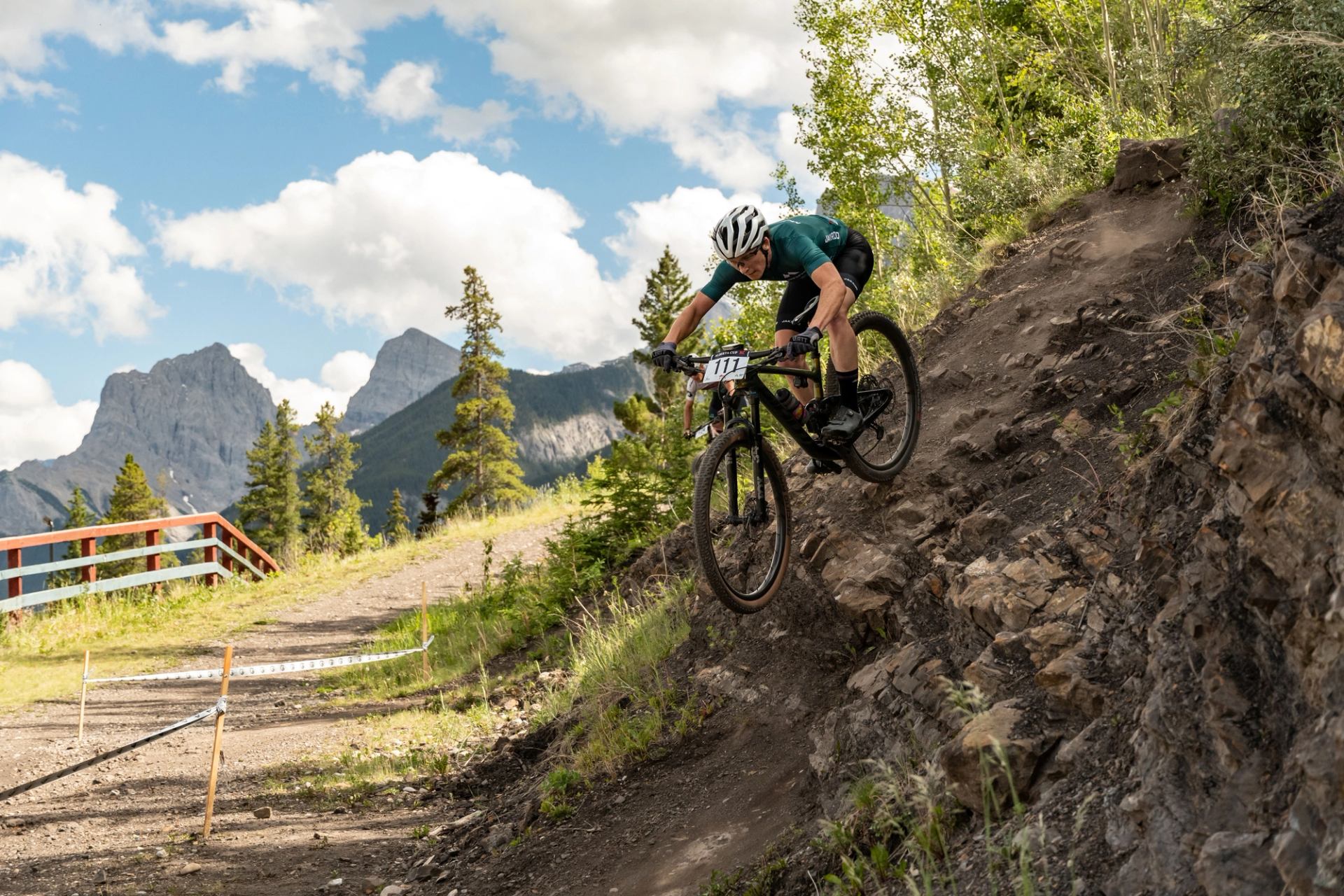 Mountain bike racer descending a steep, rocky trail with trees and mountain peaks in the background.