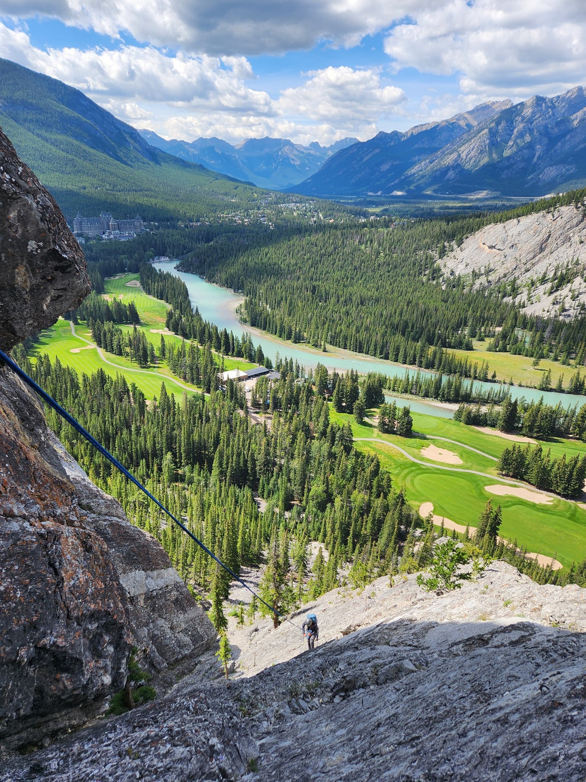 A person rappels down a rocky cliff overlooking a vibrant mountain valley featuring a turquoise river, a golf course, a large hotel, and distant peaks.