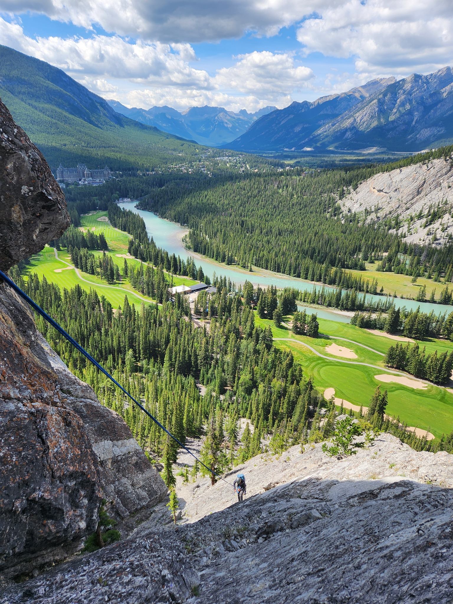 A person rappels down a rocky cliff overlooking a vibrant mountain valley featuring a turquoise river, a golf course, a large hotel, and distant peaks.