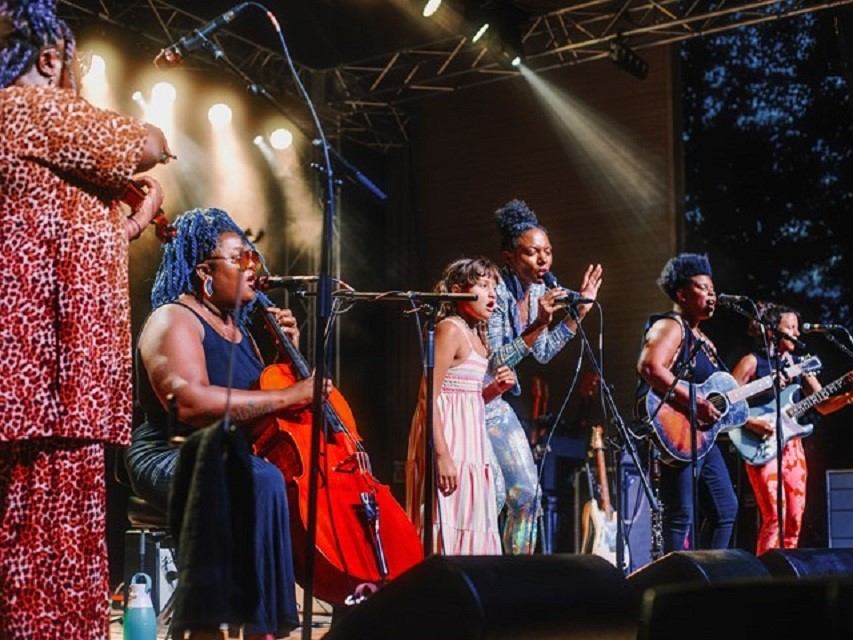 Musicians perform onstage with microphones, cello and guitars under bright concert lights.