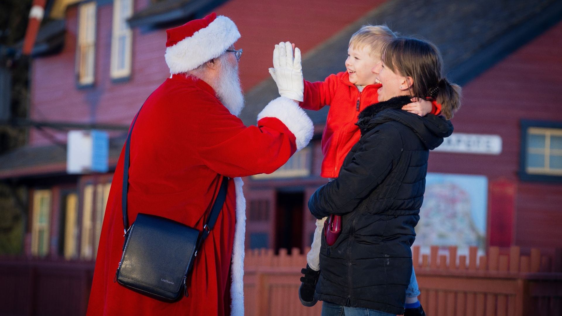 Santa gives a high-five to a child held by an adult outside a festive red building.