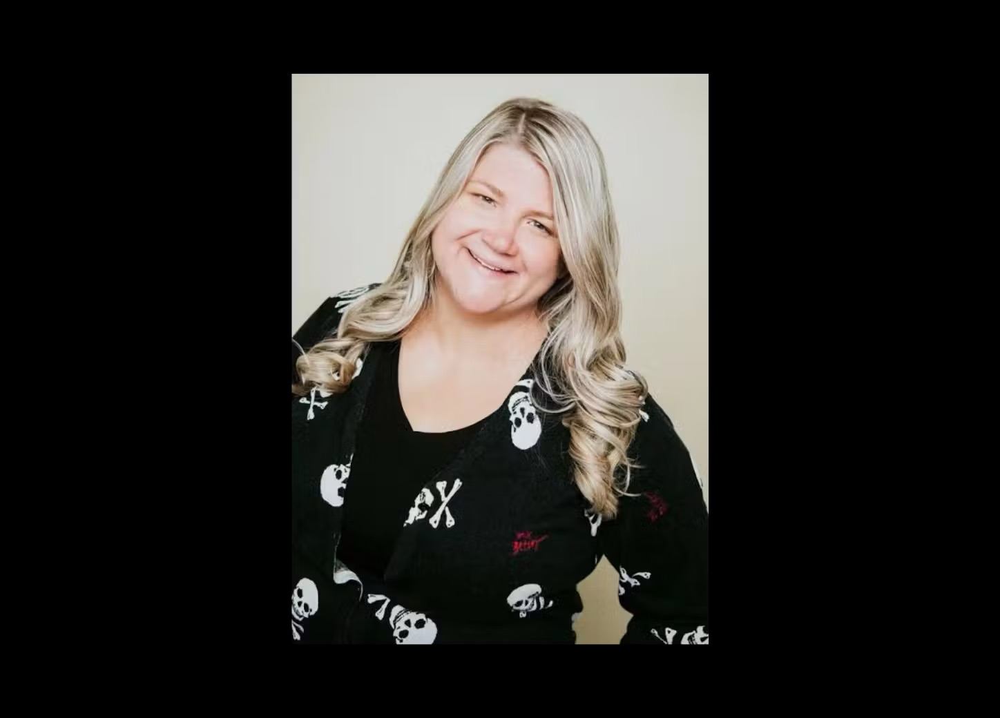 Woman with long blonde hair smiling in a black patterned shirt portrait against a light background.