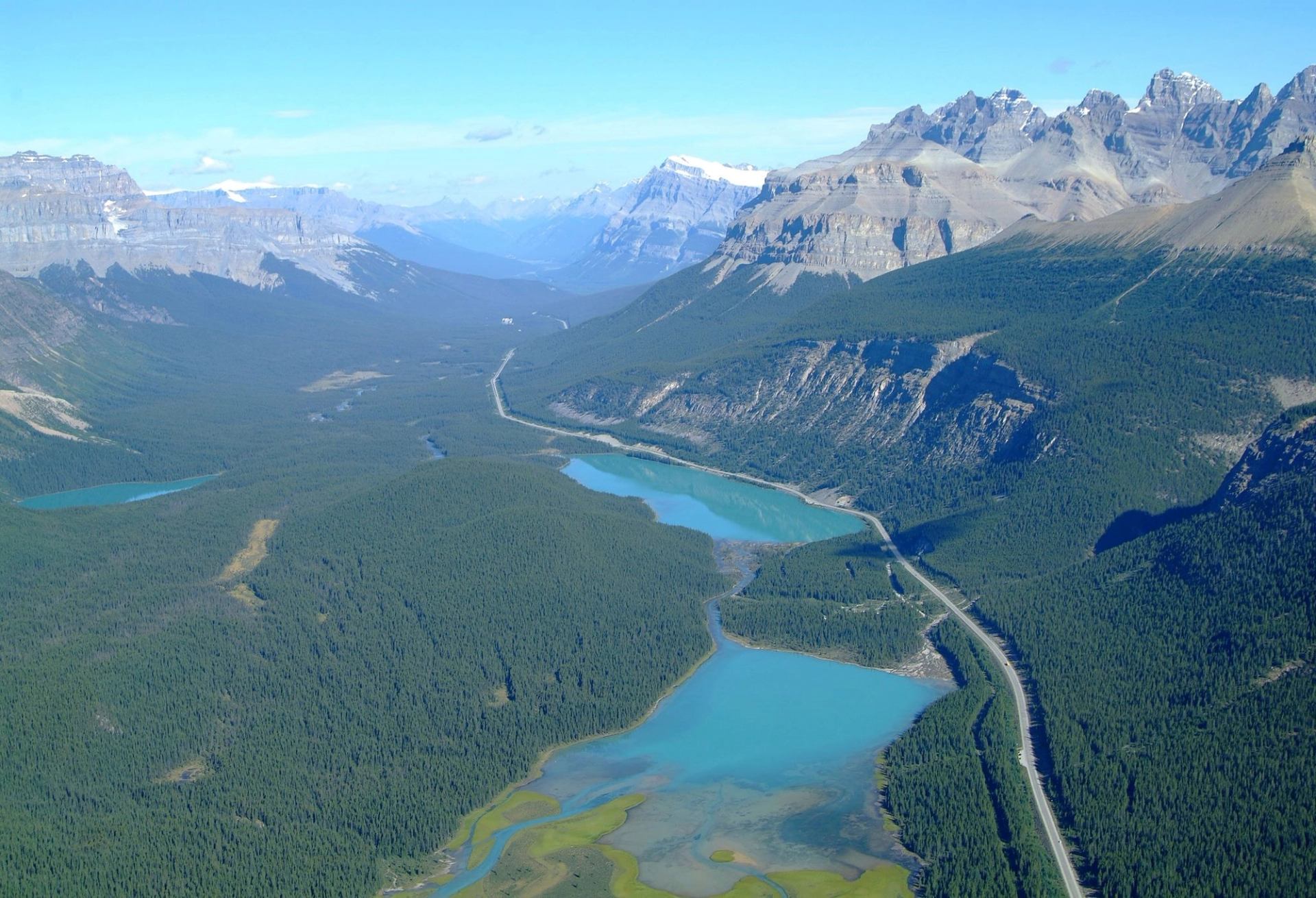 Aerial view of a winding road and turquoise rivers flanked by lush forests and rugged mountains under a clear blue sky, conveying serenity and grandeur.