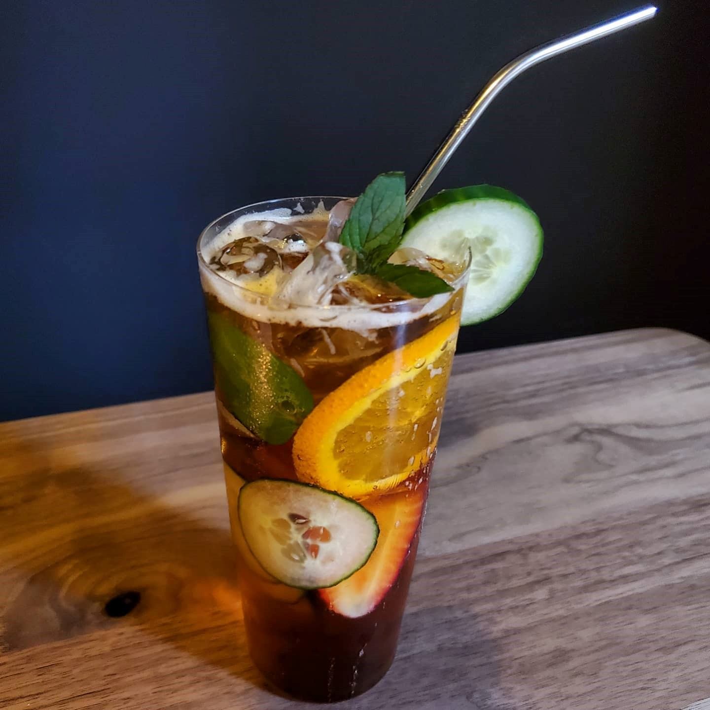 Tall glass with dark drink, ice, fruit slices, mint, and metal straw on wood surface.