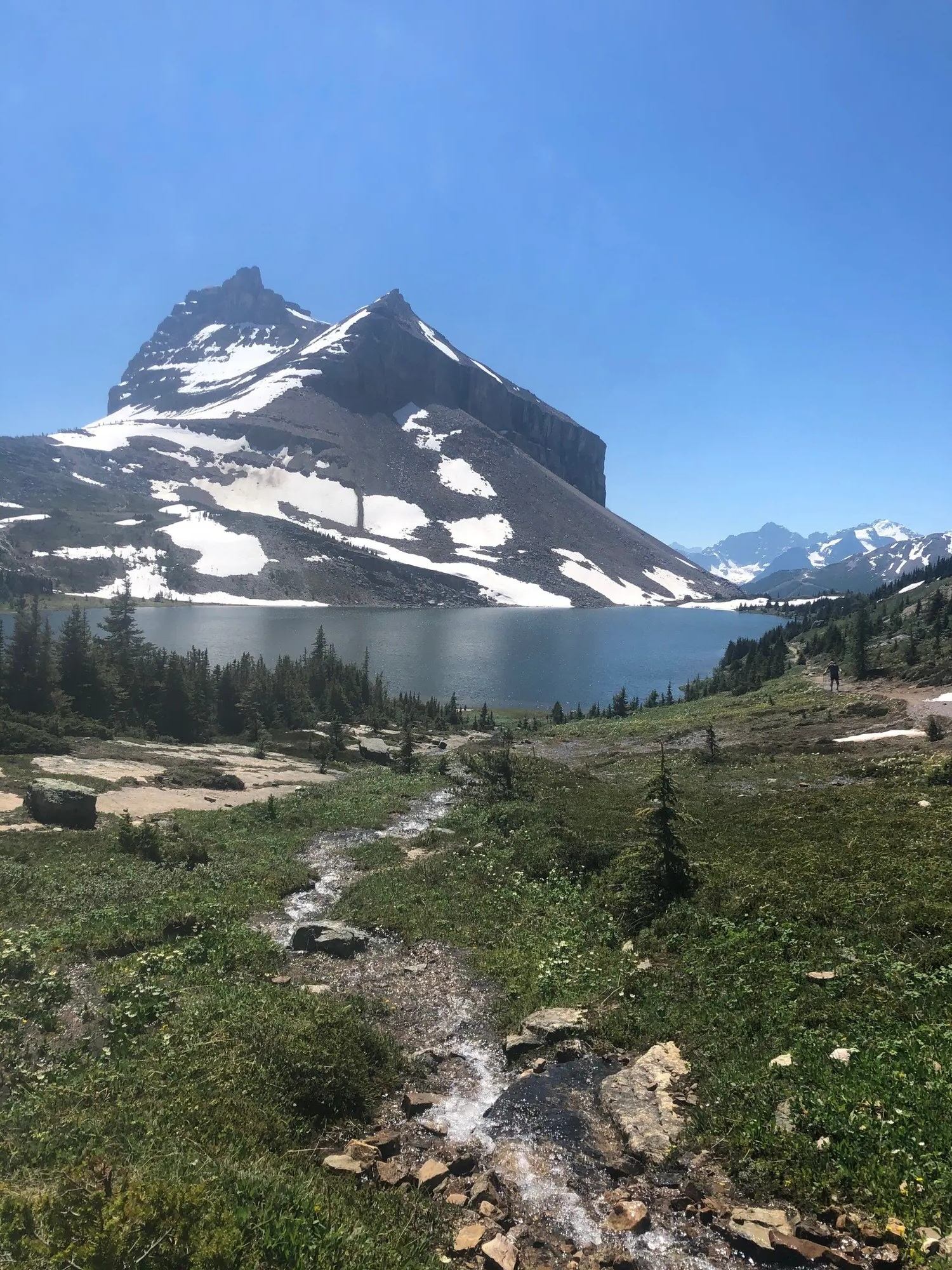 Mountain stream flows toward a lake beneath snow-covered peaks and a clear blue sky.