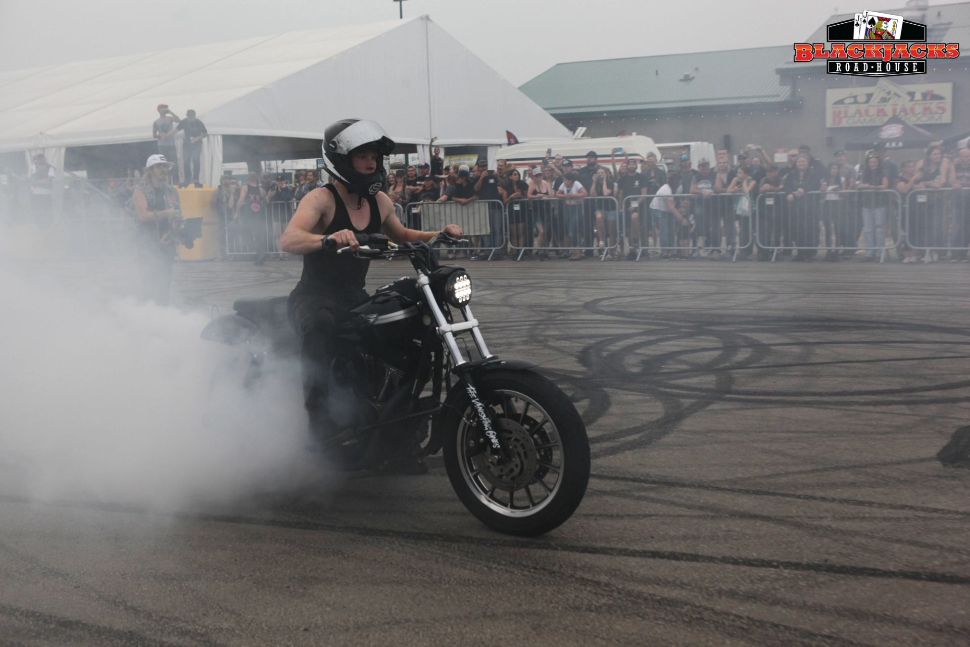 Motorcycle rider performs stunt on black motorcycle with smoke, audience seated behind barriers at B.
