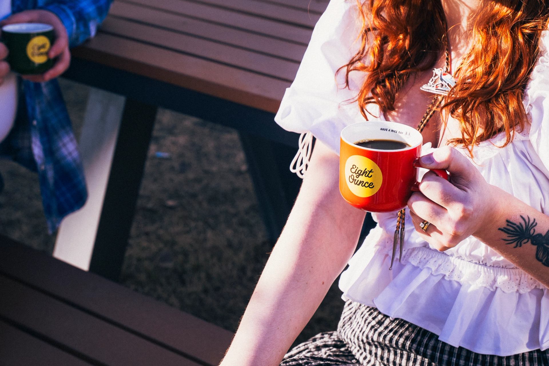Close‑up of a person holding a red coffee mug while sitting outdoors at a picnic table.