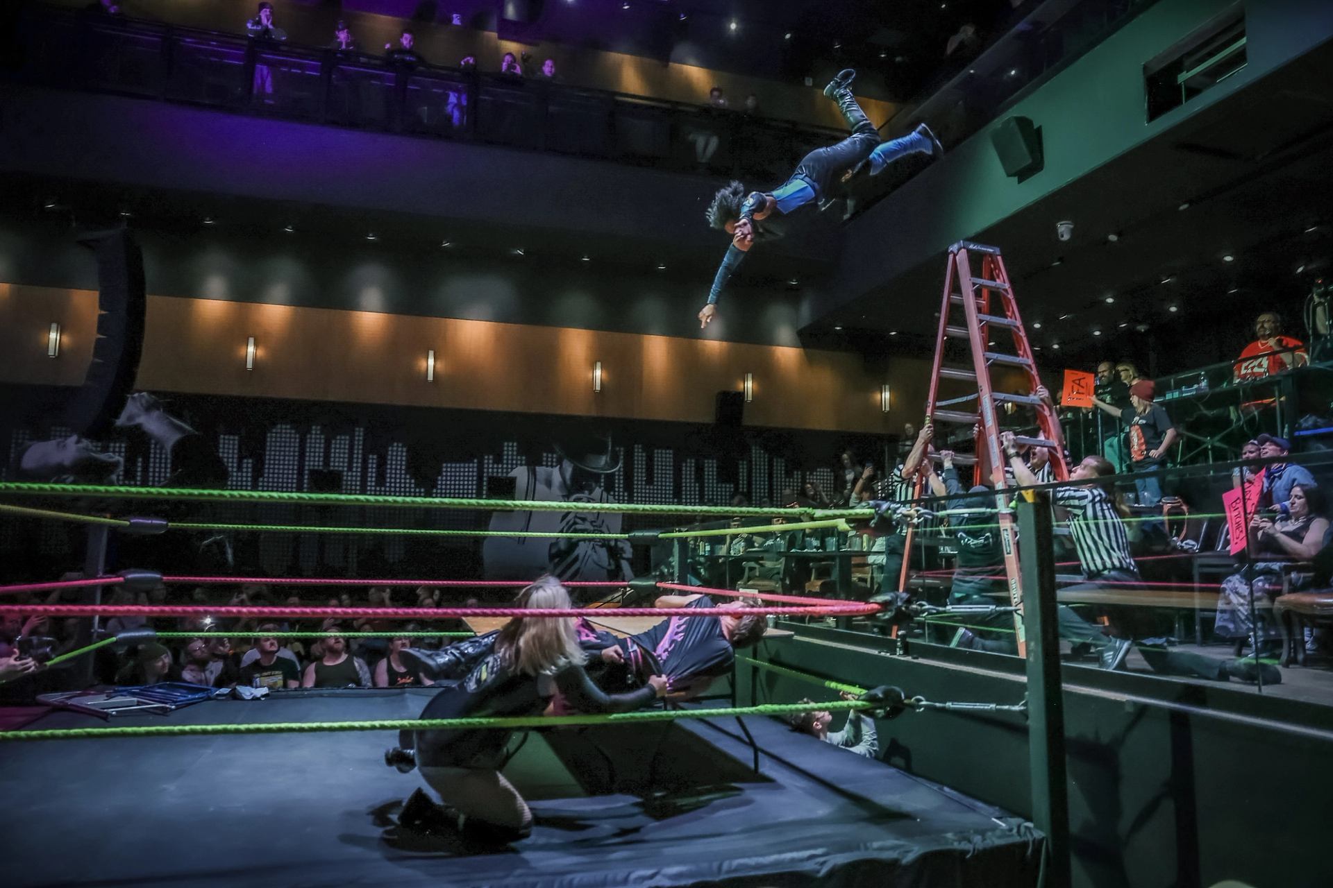 Wrestler dives from a ladder toward the ring as spectators watch from the balcony.
