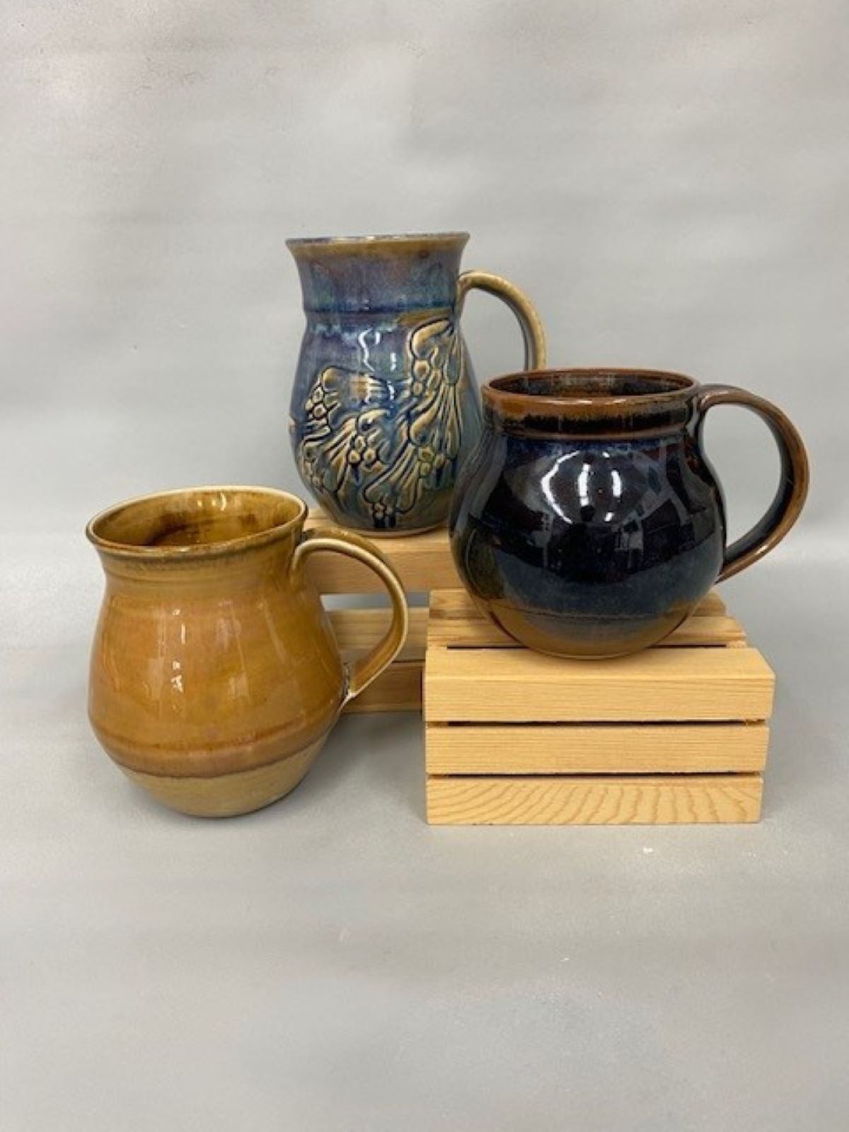 Three handmade ceramic vessels with blue and golden glazes, arranged on wooden blocks against a neutral backdrop.