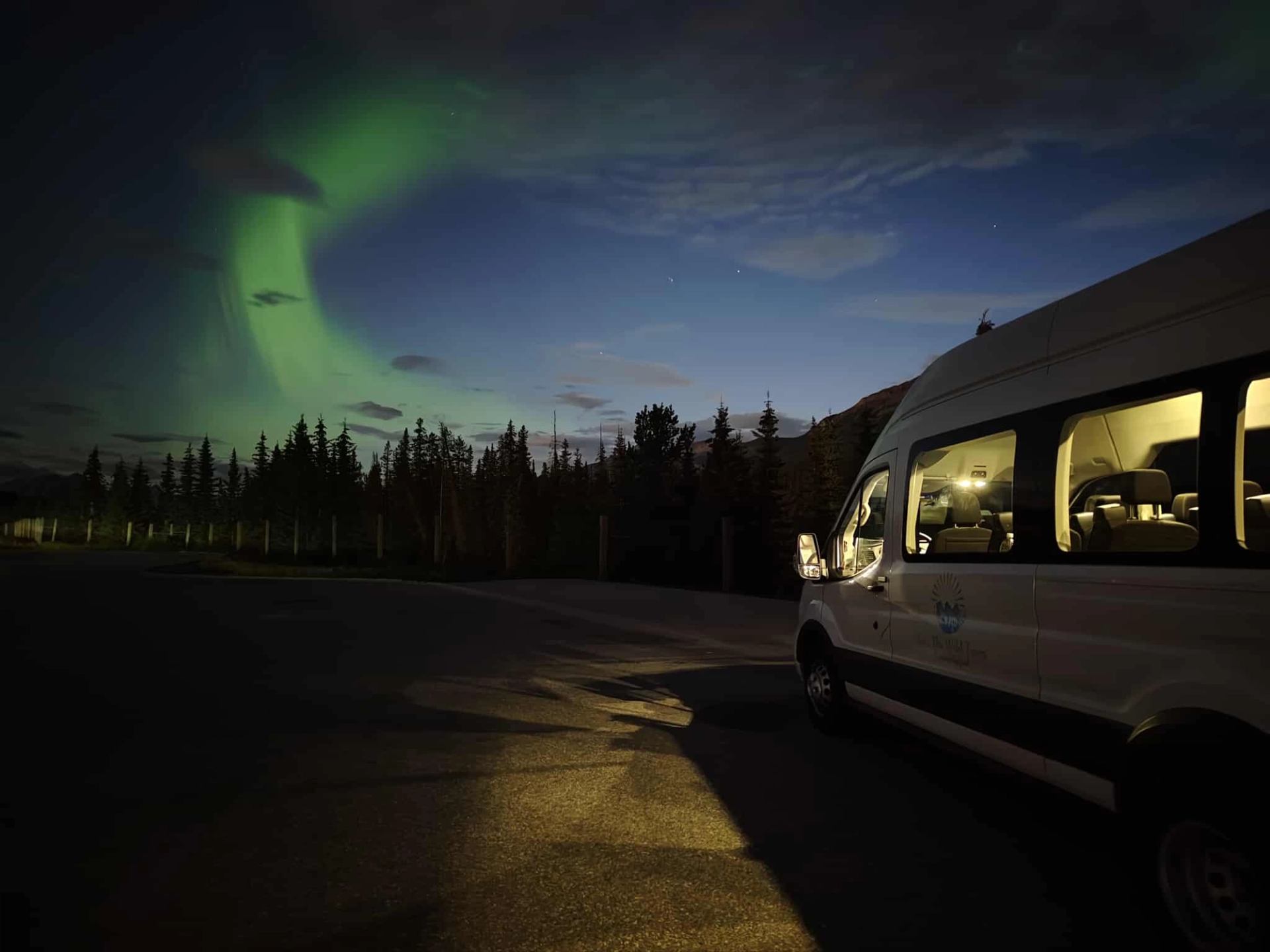 A van parked under a night sky with green aurora above a treeline.